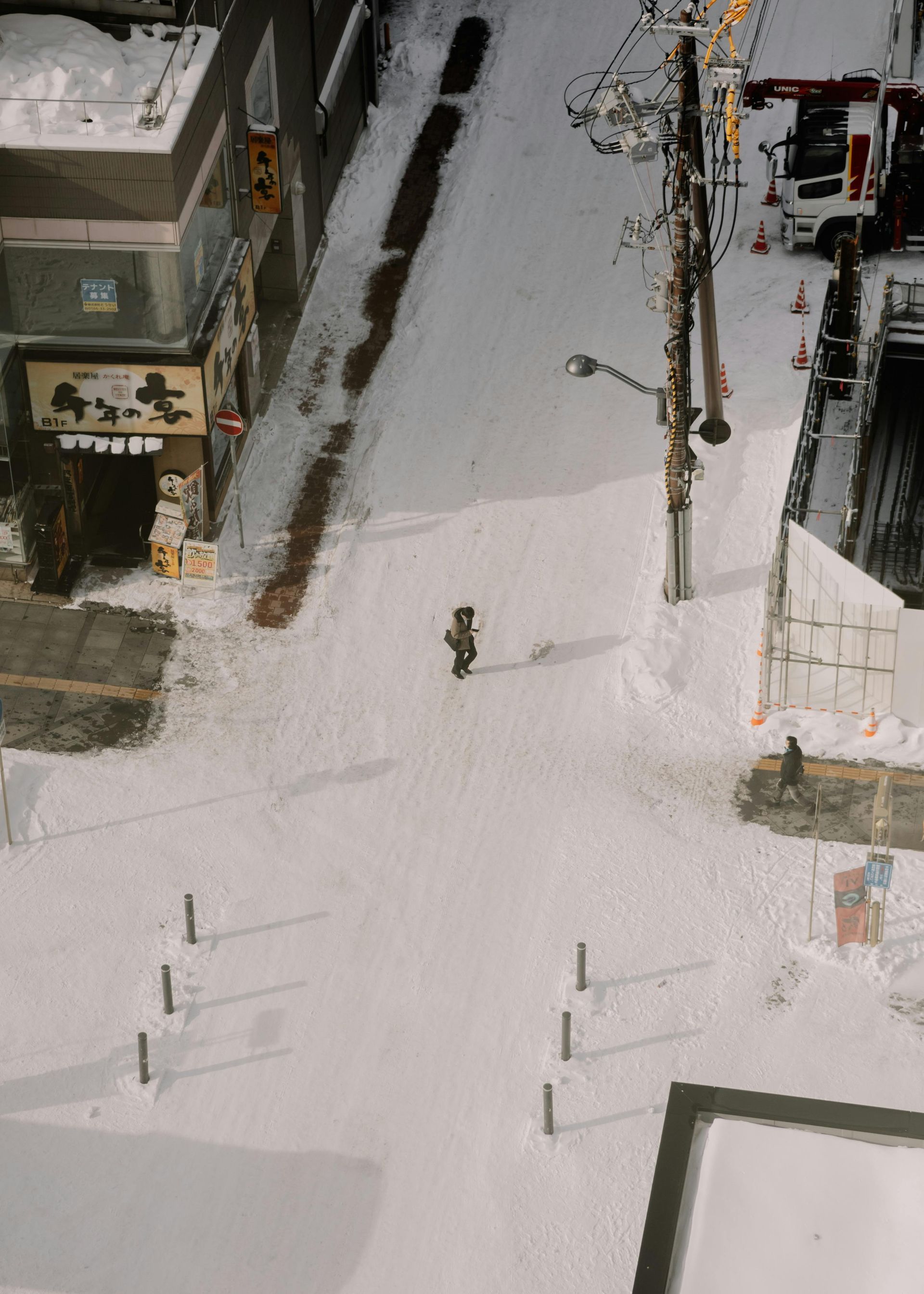 Snowy street scene, a person walking down a path through deep snow. Buildings and a utility pole line the street.