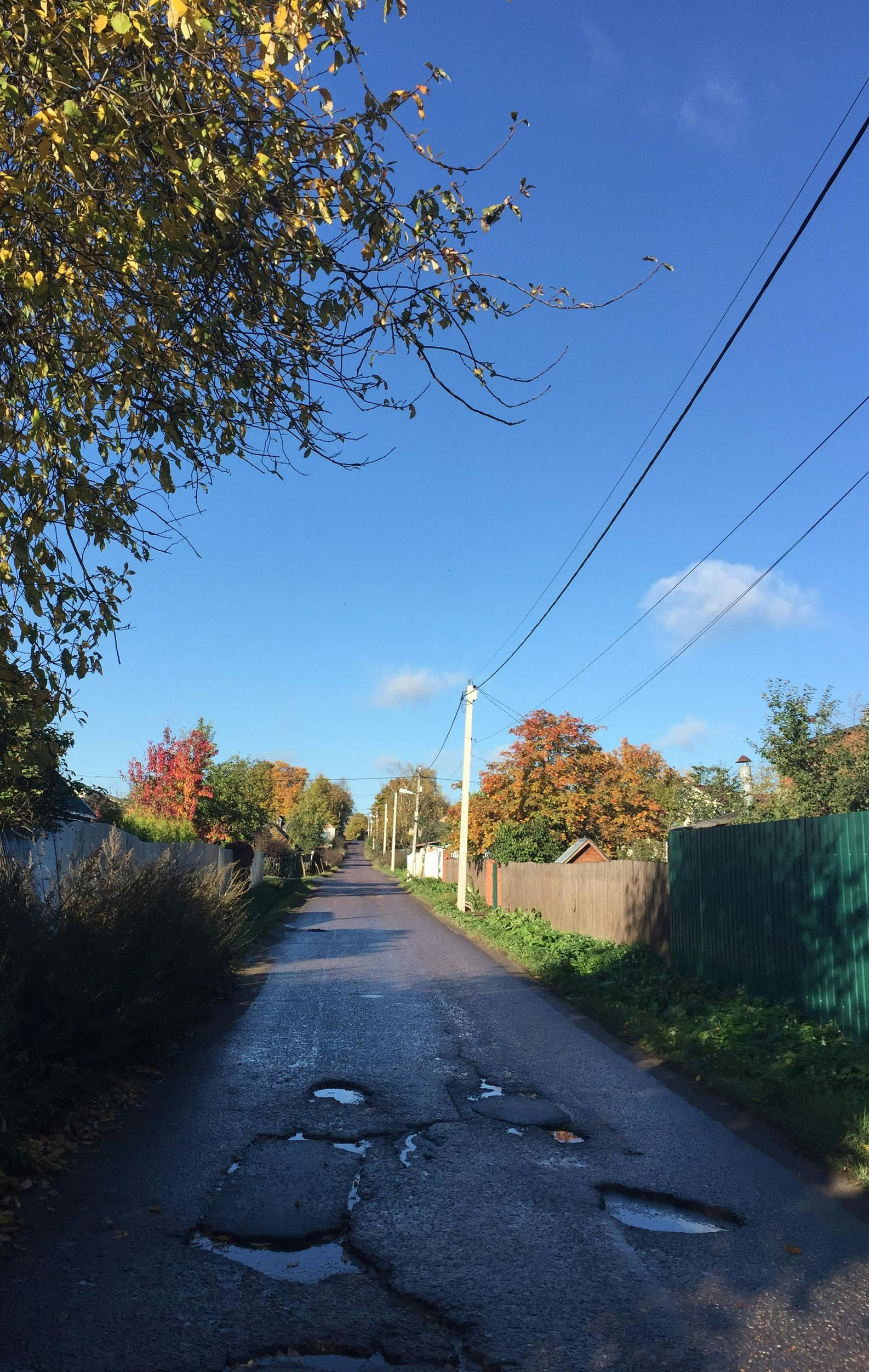 Paved road with potholes in a rural area, lined with houses, trees with autumn colors, under a blue sky.