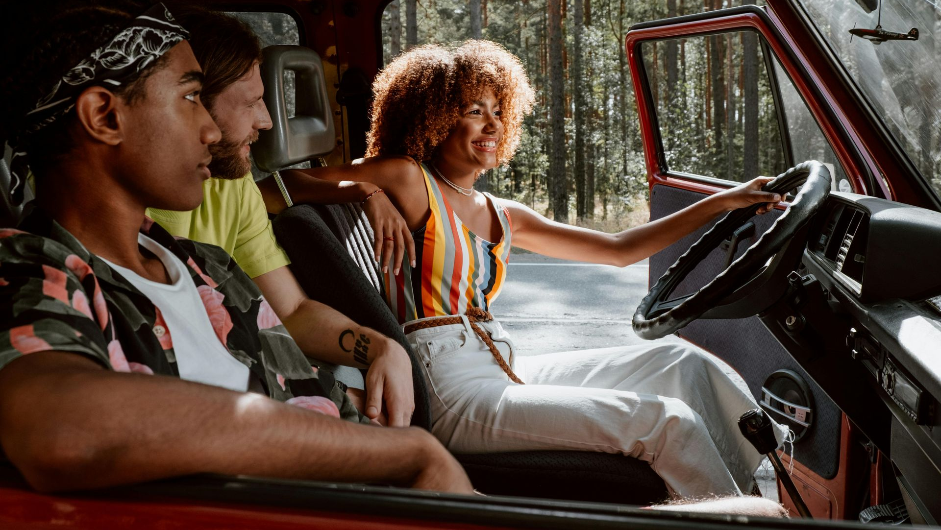 Three people sit in a red van, one driving, with sunlight streaming through the windows.
