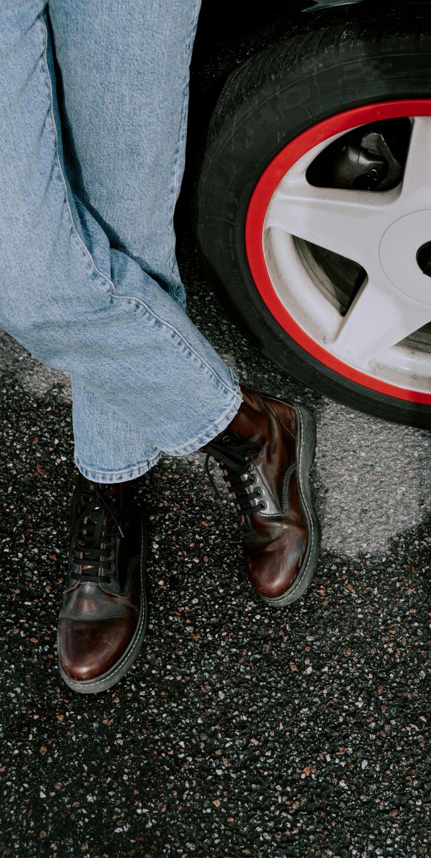 Person in blue jeans and brown boots leaning against a car wheel with a red rim.