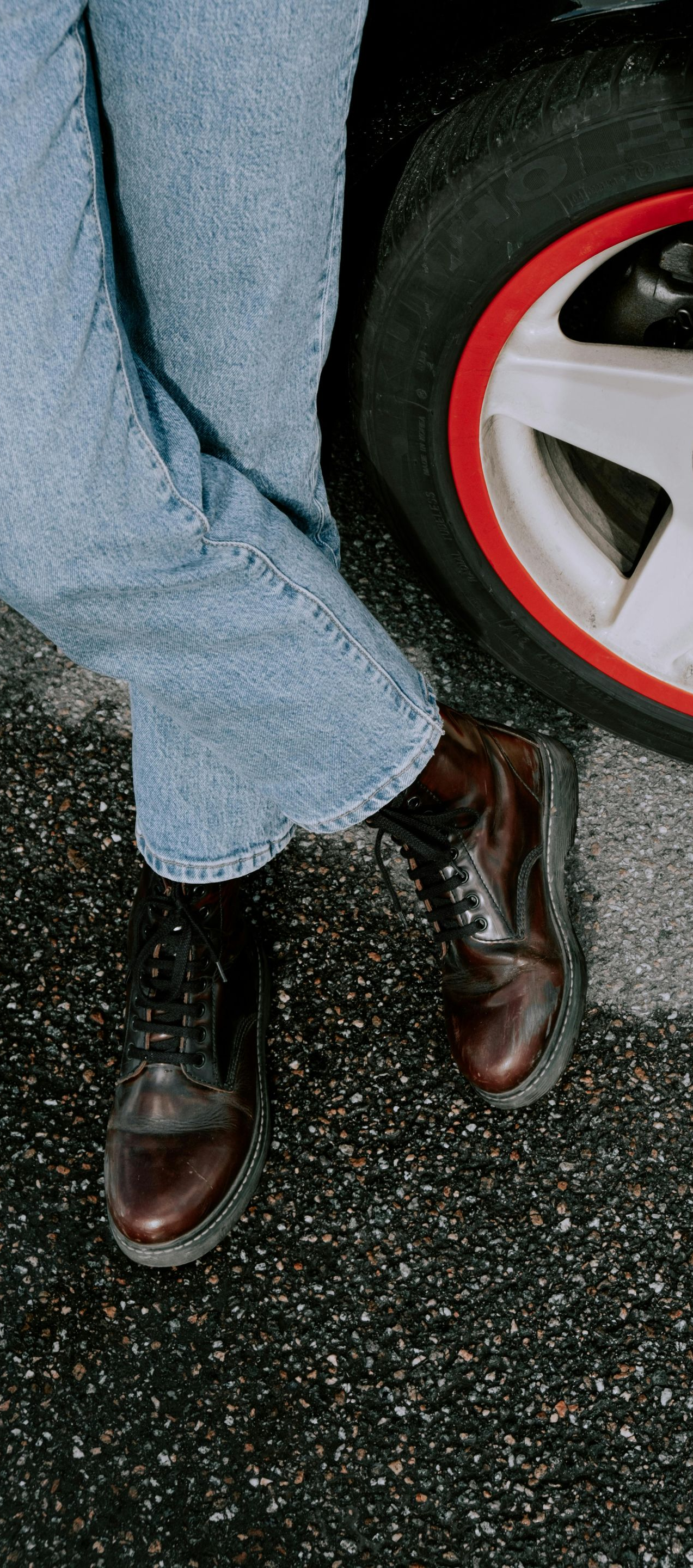 Person in blue jeans and brown boots leaning against a car wheel with a red rim.