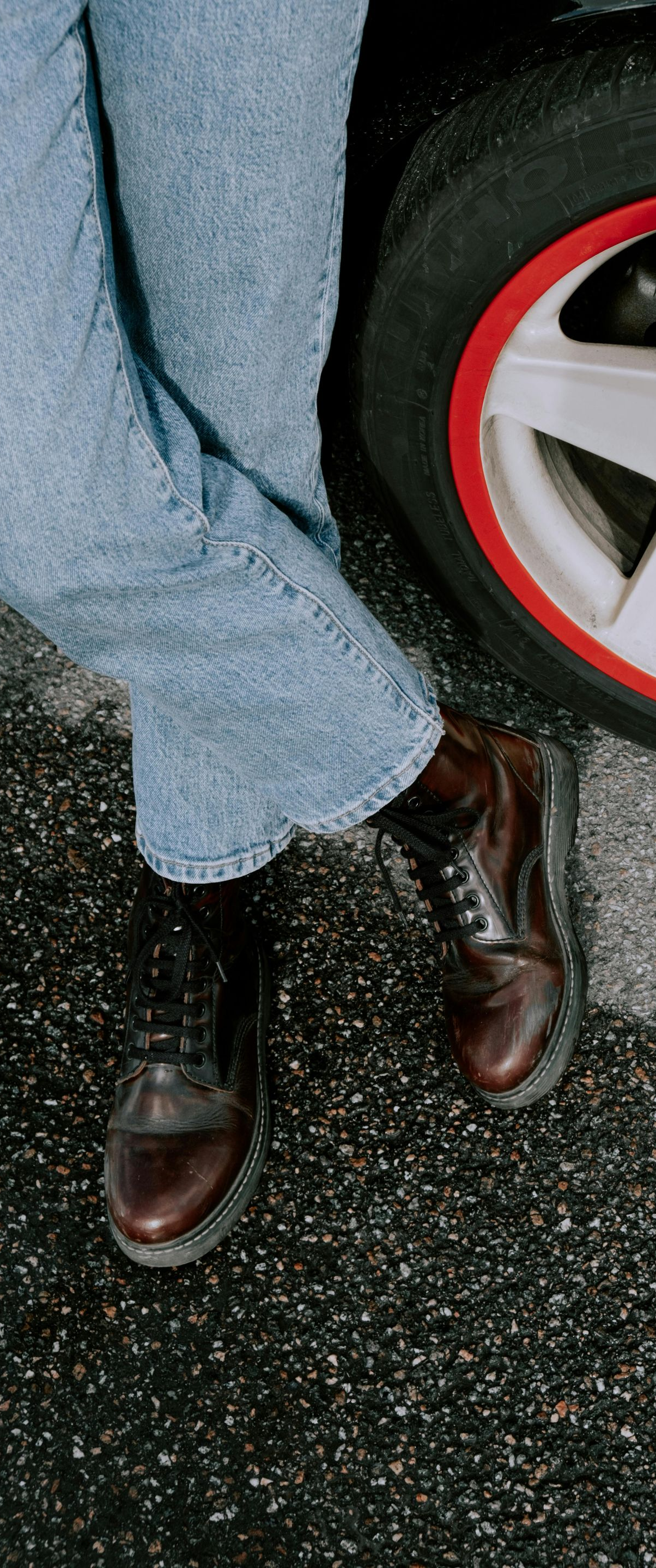 Person in blue jeans and brown boots leaning against a car wheel with a red rim.