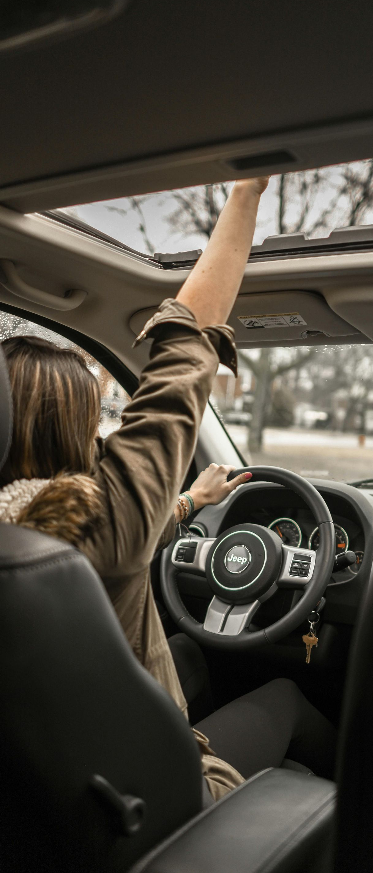 A person in a car reaching for the sunroof, while driving with the other hand on the steering wheel.