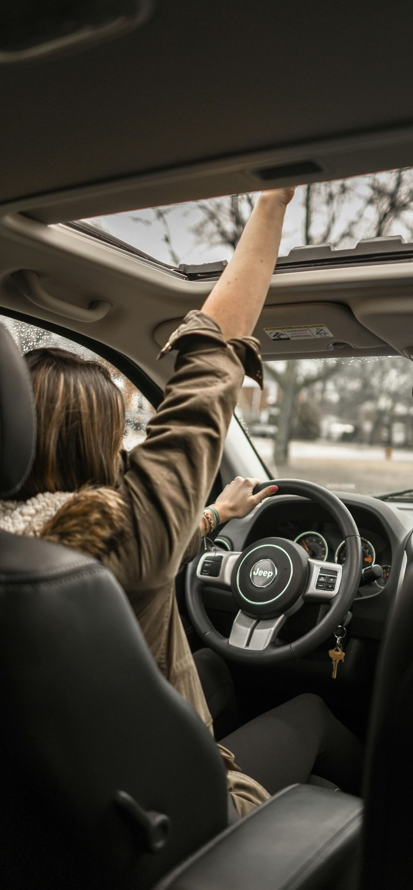 A person in a car reaching for the sunroof, while driving with the other hand on the steering wheel.