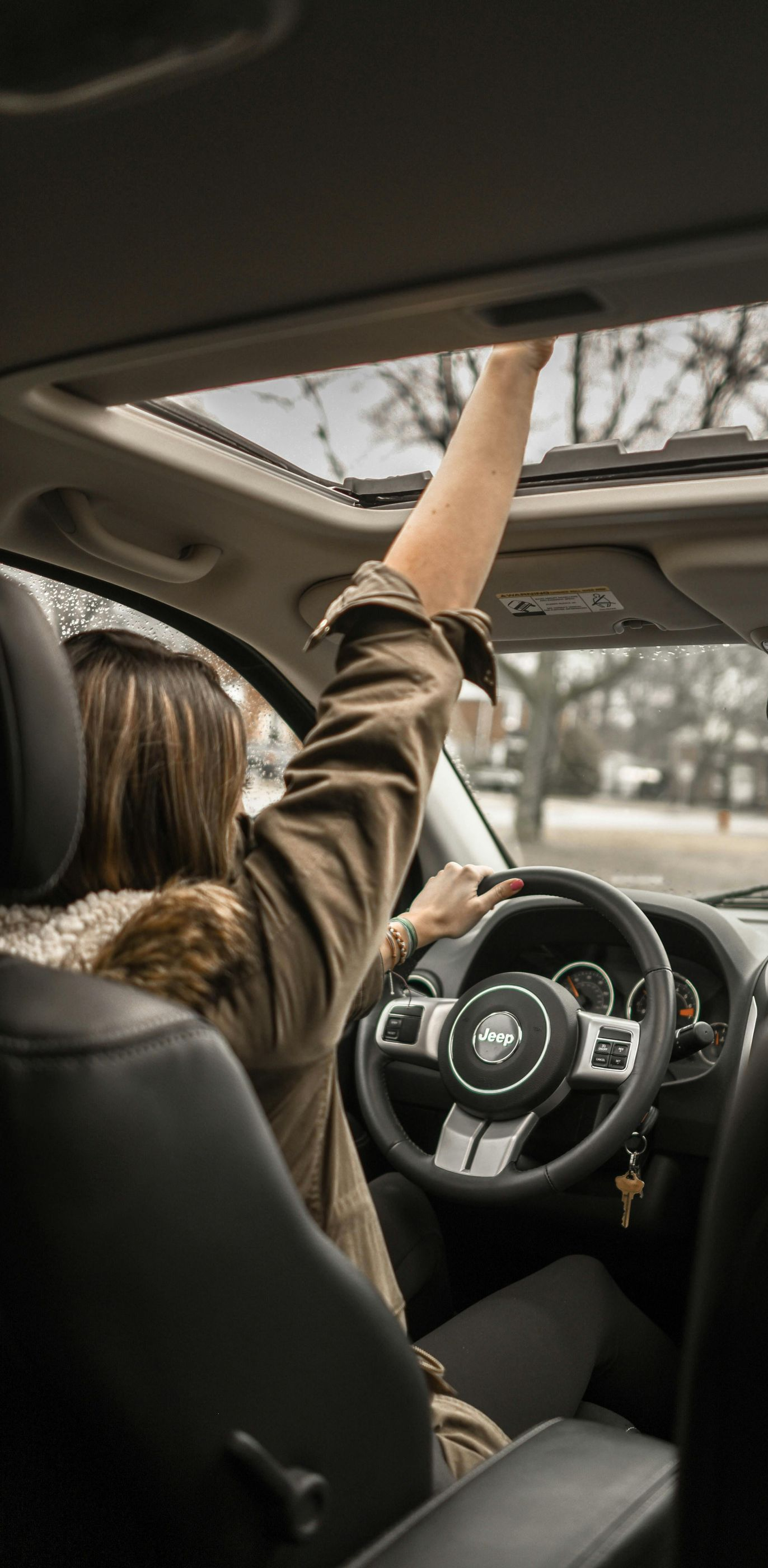 A person in a car reaching for the sunroof, while driving with the other hand on the steering wheel.