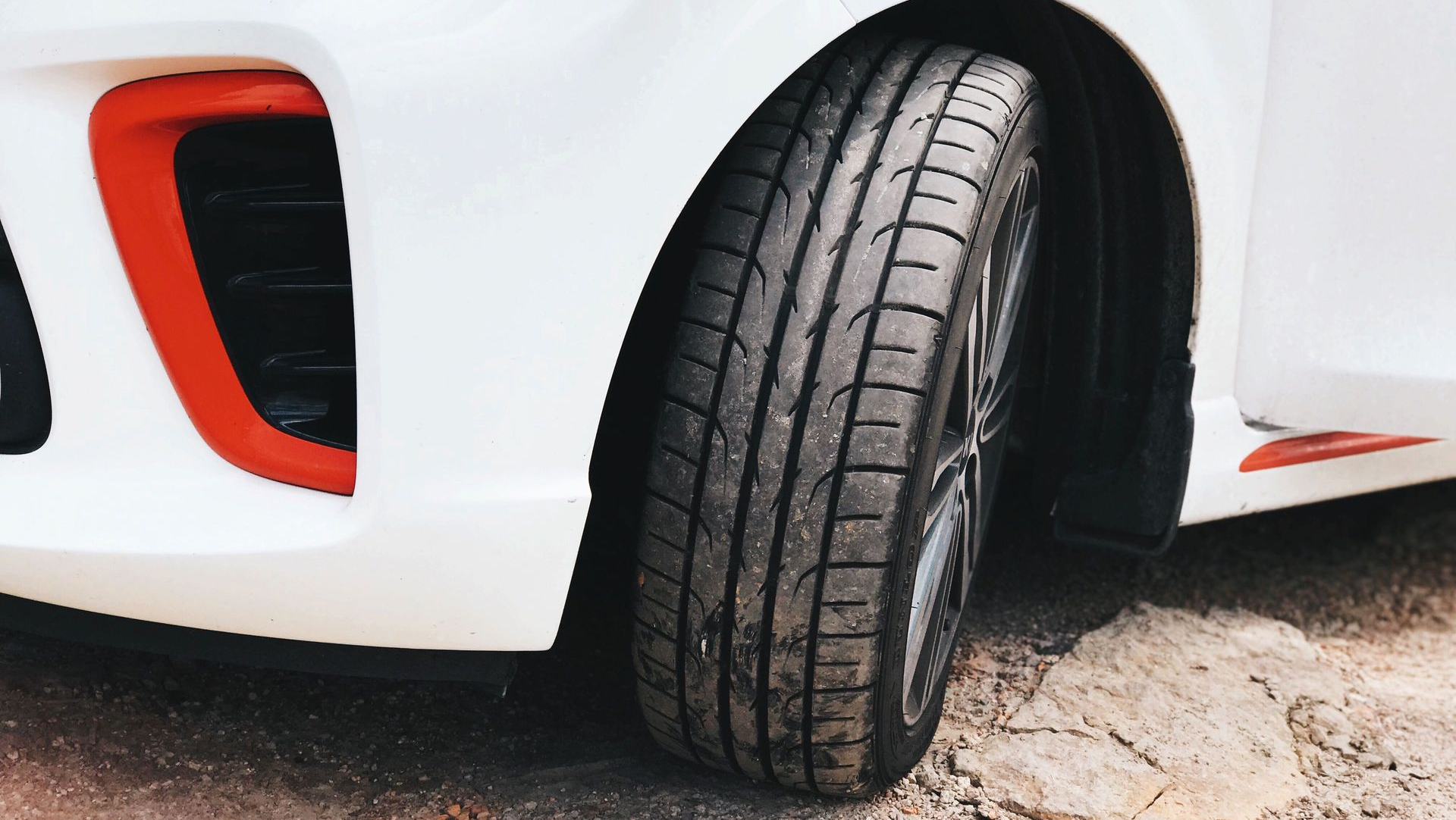 Close-up of a white car’s front wheel on rough pavement, with red trim near the bumper.