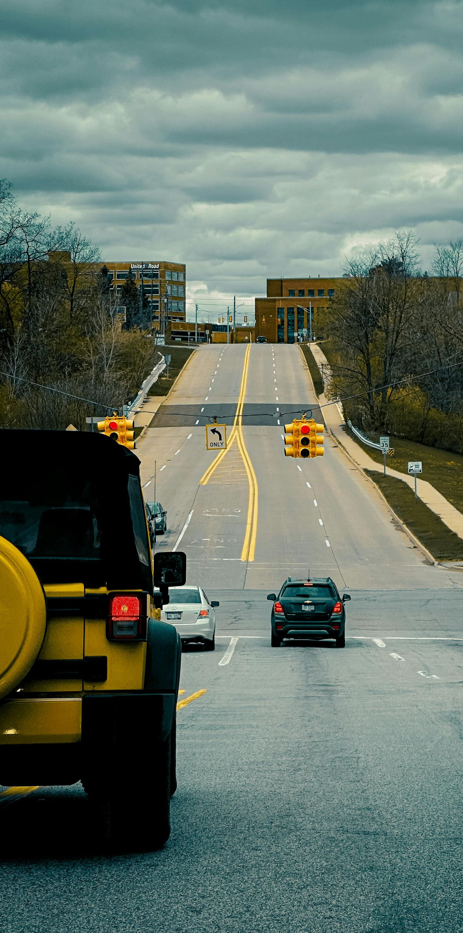 Cars driving down a wet road toward a hill under cloudy skies, with trees lining both sides.