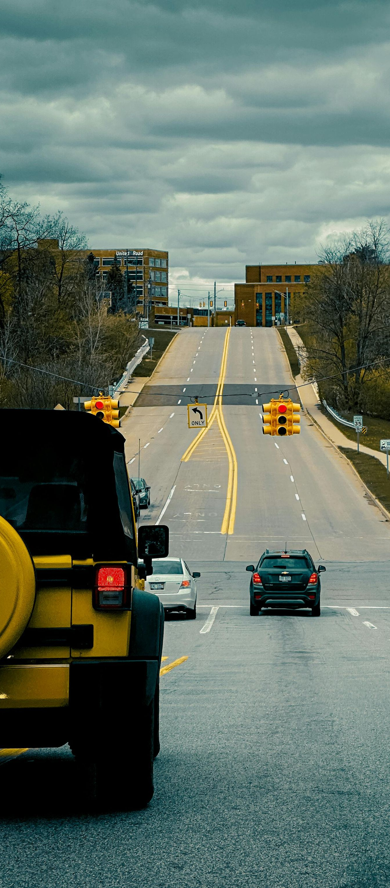 Cars driving down a wet road toward a hill under cloudy skies, with trees lining both sides.