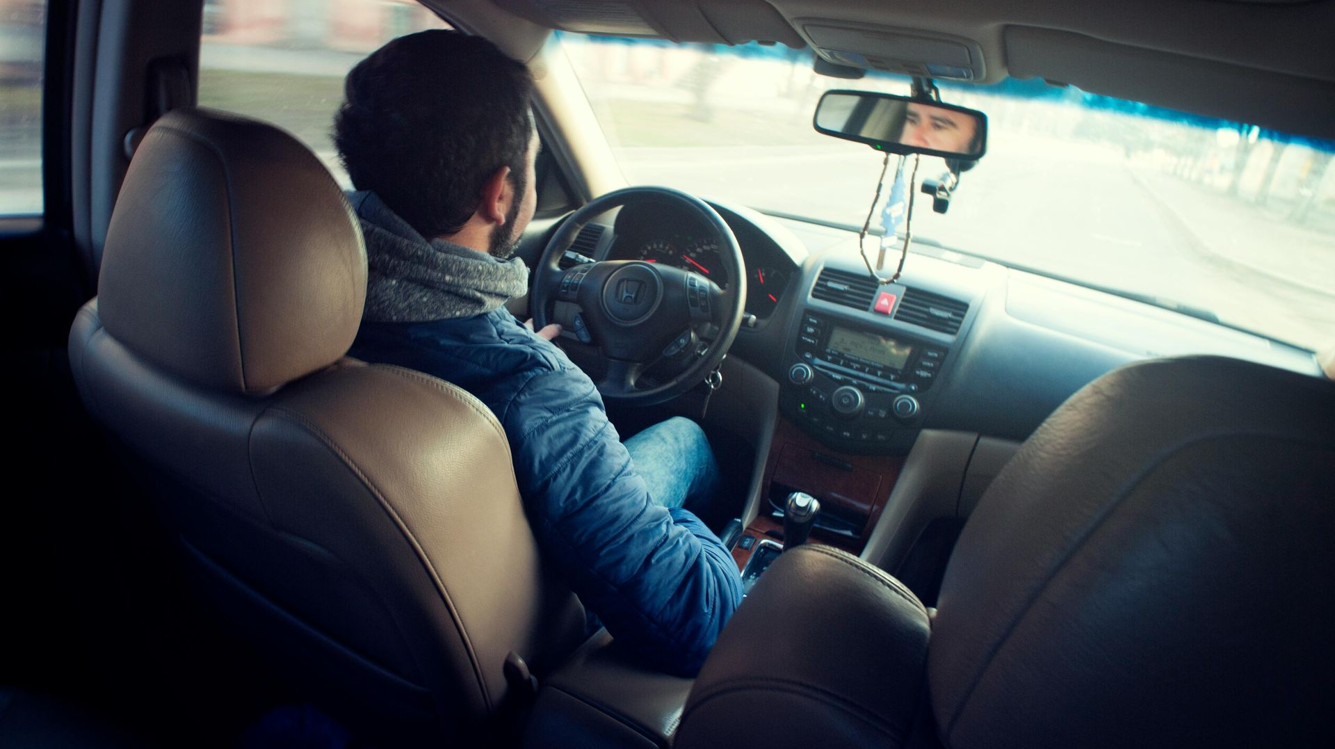 Man driving a car, view from back seat. He wears a blue jacket and looks forward. Interior of a car.