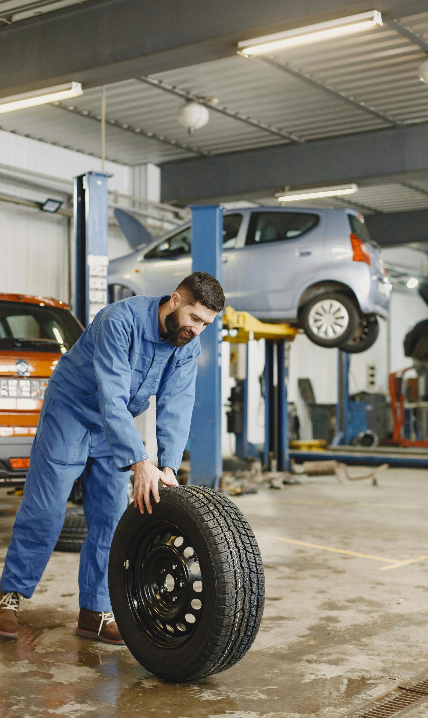 Mechanic in blue overalls rolls a tire across a garage floor. Car is raised on a lift in the background.