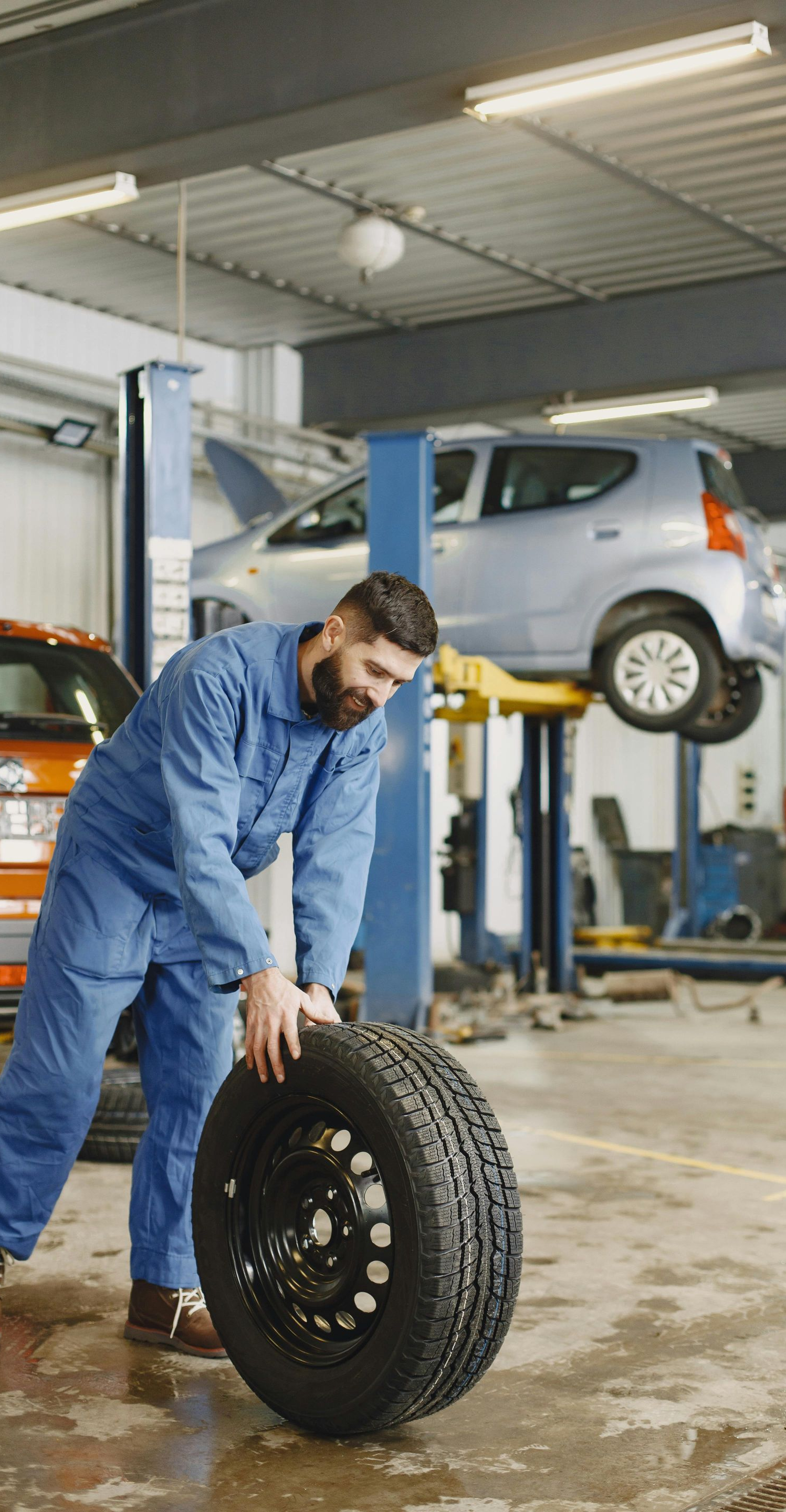 Mechanic in blue overalls rolls a tire across a garage floor. Car is raised on a lift in the background.