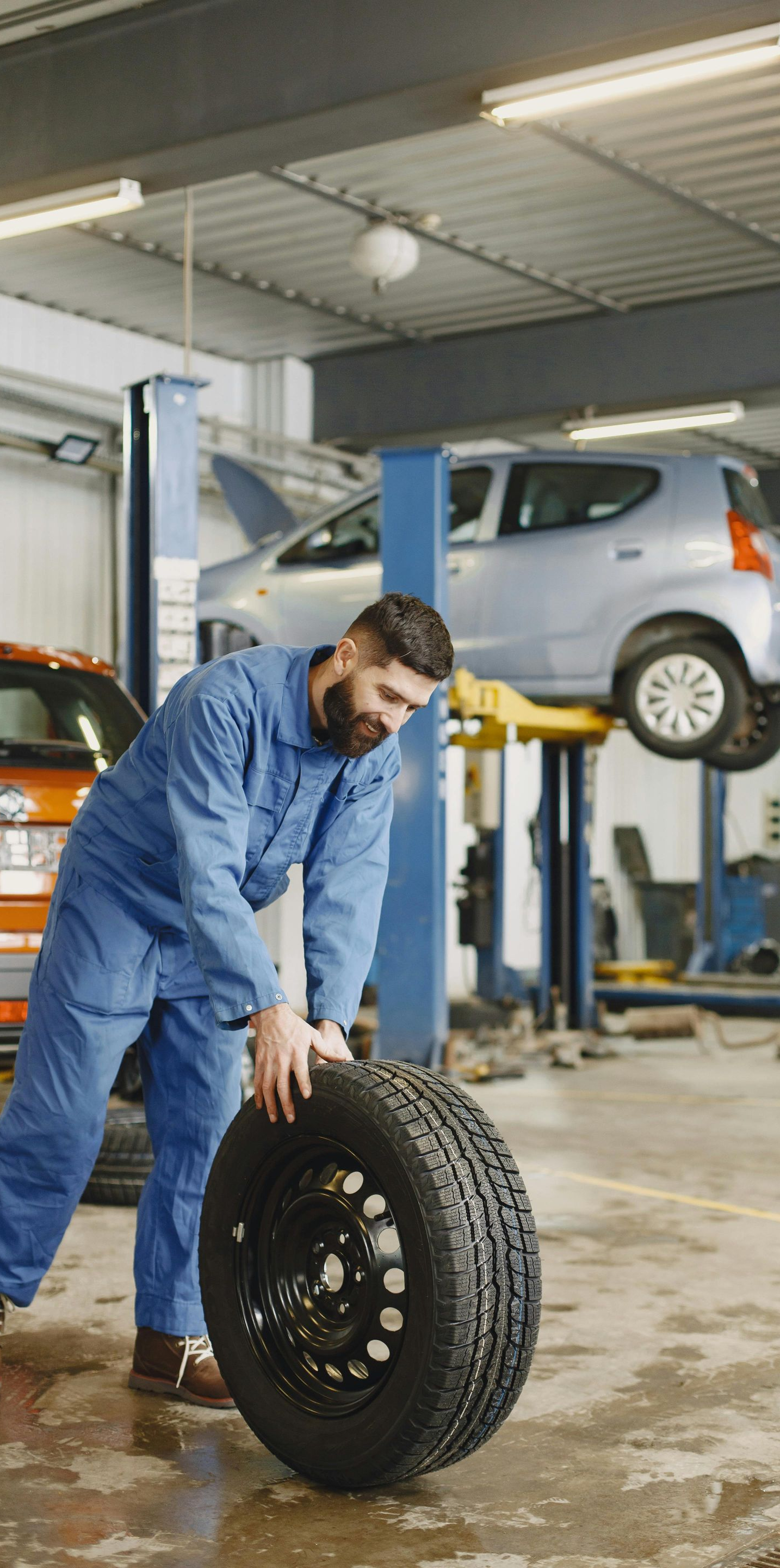 Mechanic in blue overalls rolls a tire across a garage floor. Car is raised on a lift in the background.