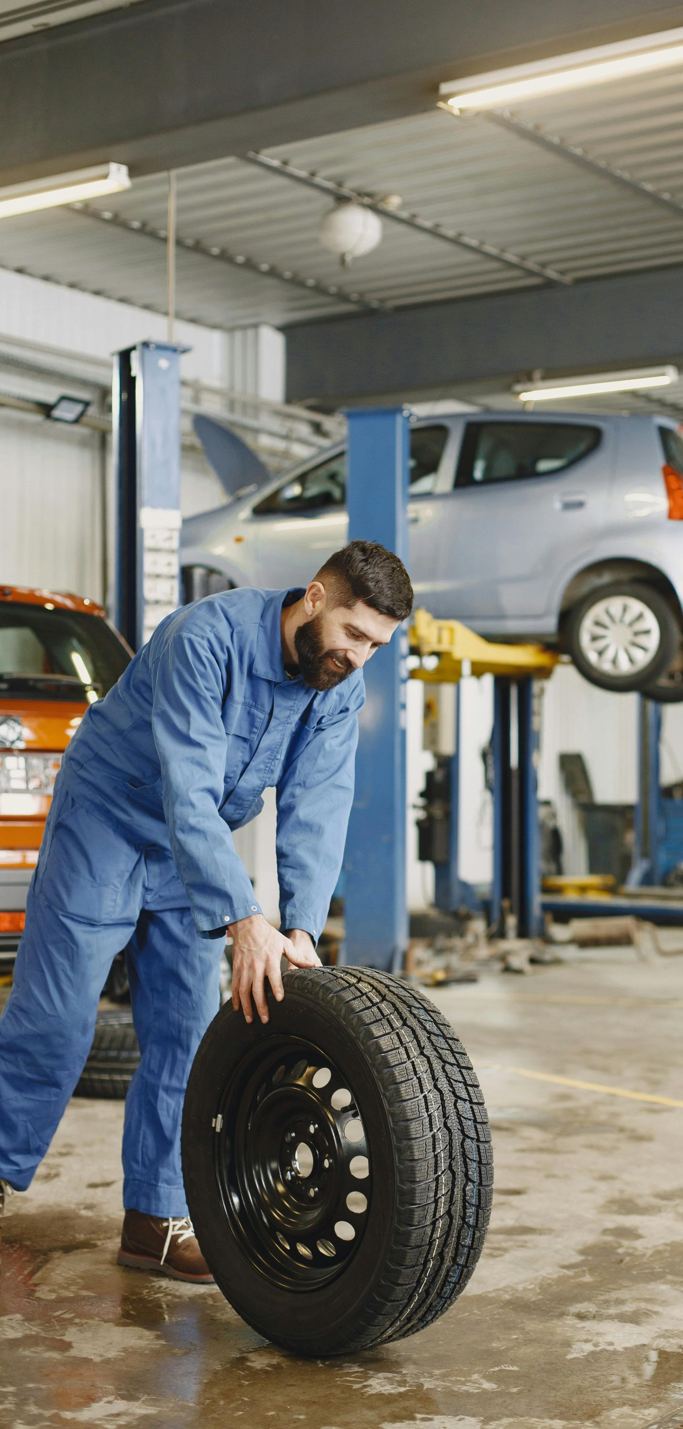 Mechanic in blue overalls rolls a tire across a garage floor. Car is raised on a lift in the background.
