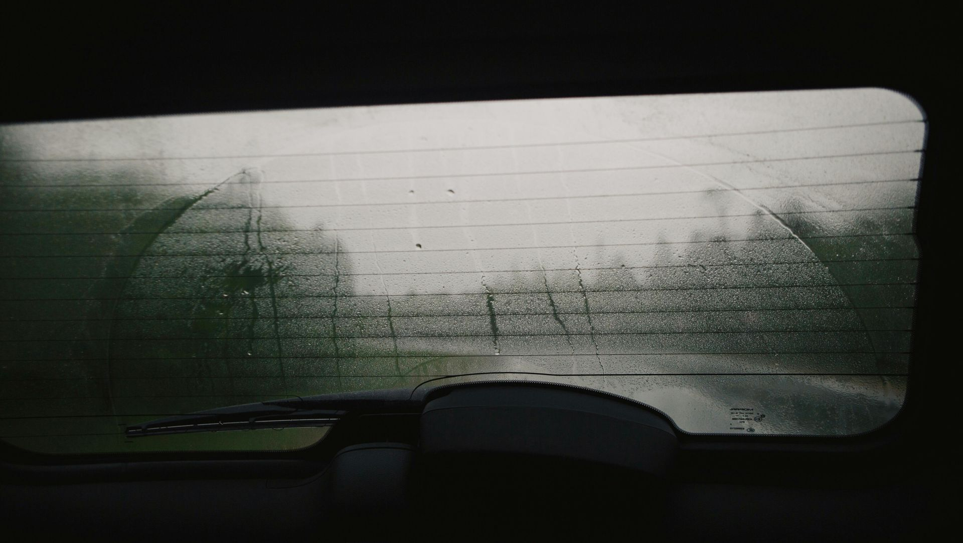 Rear car window obscured by rain and condensation; wipers clearing view of green landscape.
