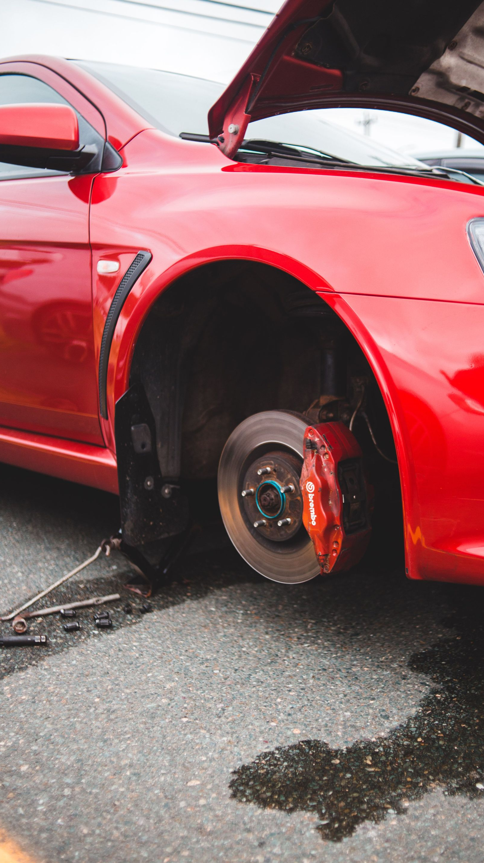 Red sports car with hood open and front wheel removed, exposing the brake rotor on a wet street.