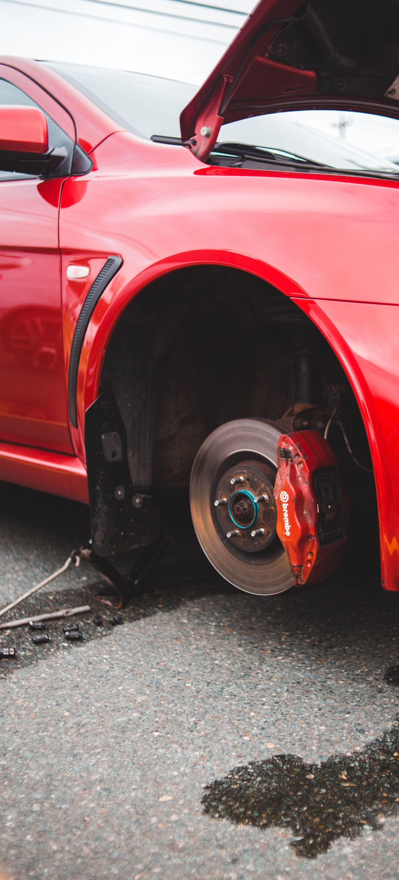 Red sports car with hood open and front wheel removed, exposing the brake rotor on a wet street.
