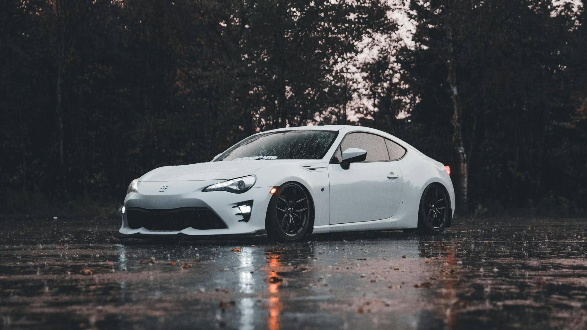 White sports car parked in rain, dark background of trees.