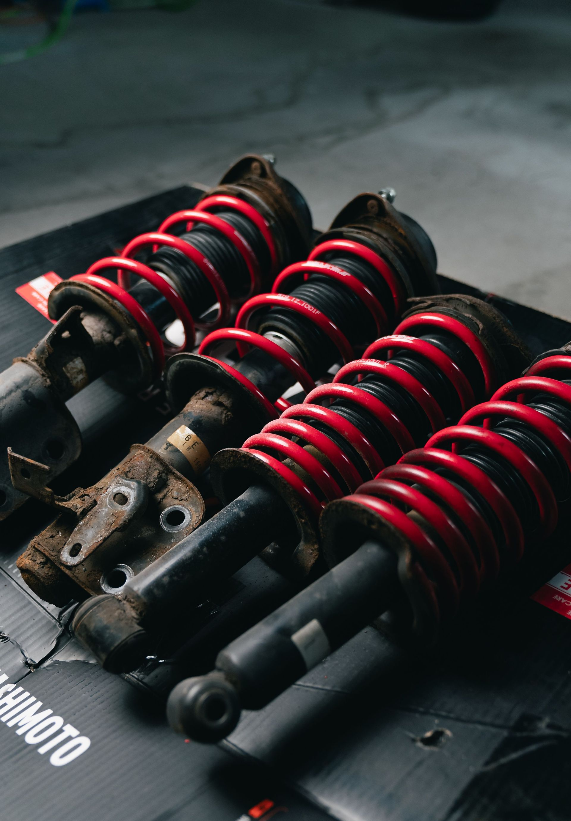 Black suspension coilover shocks with red springs arranged on a mat, close-up on a workshop floor