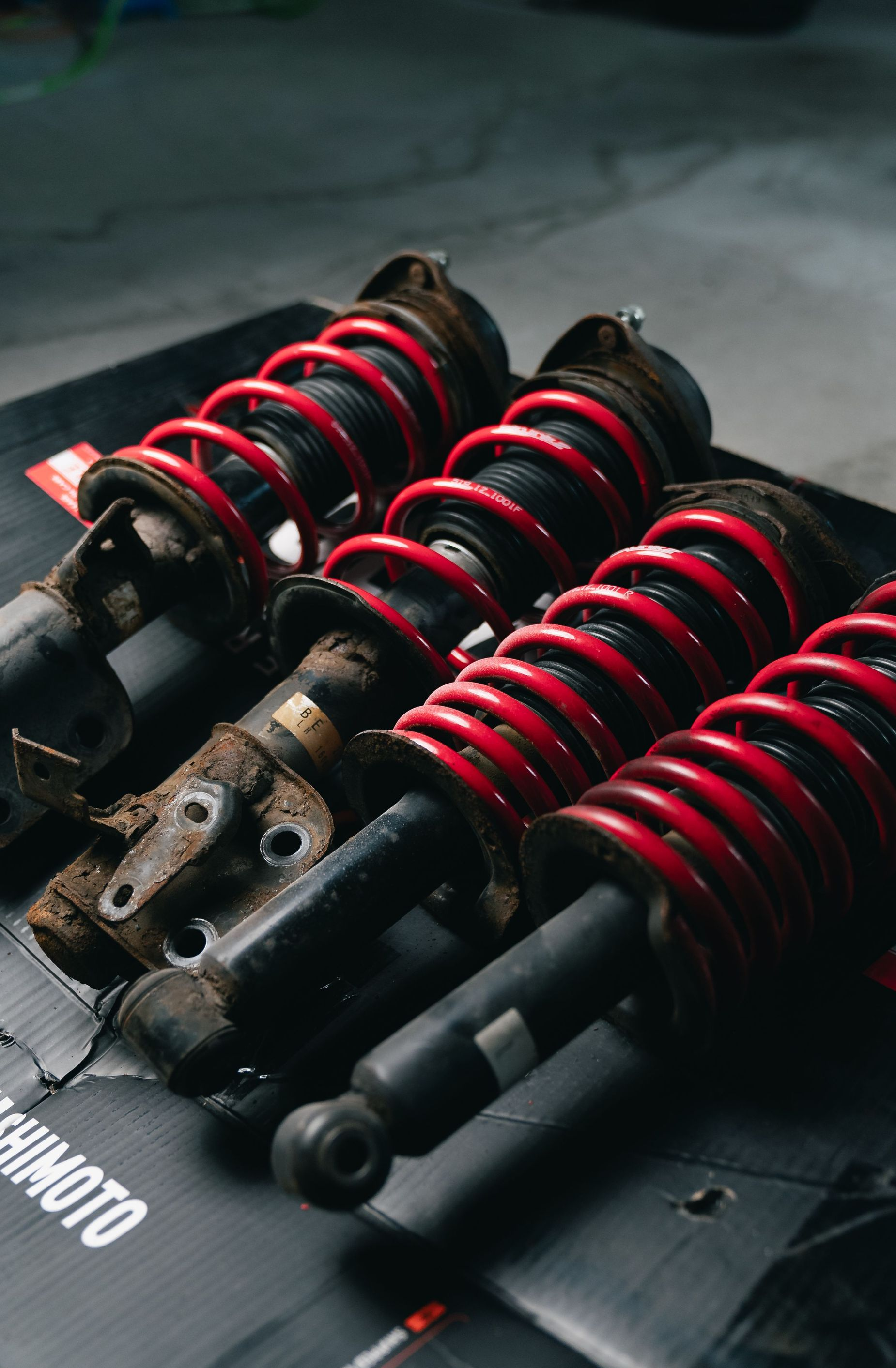 Black suspension coilover shocks with red springs arranged on a mat, close-up on a workshop floor