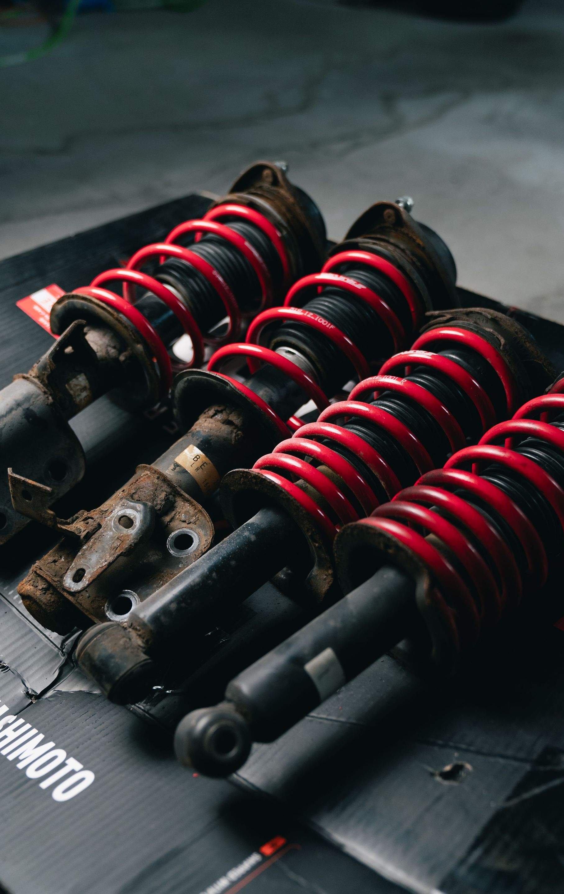 Black suspension coilover shocks with red springs arranged on a mat, close-up on a workshop floor