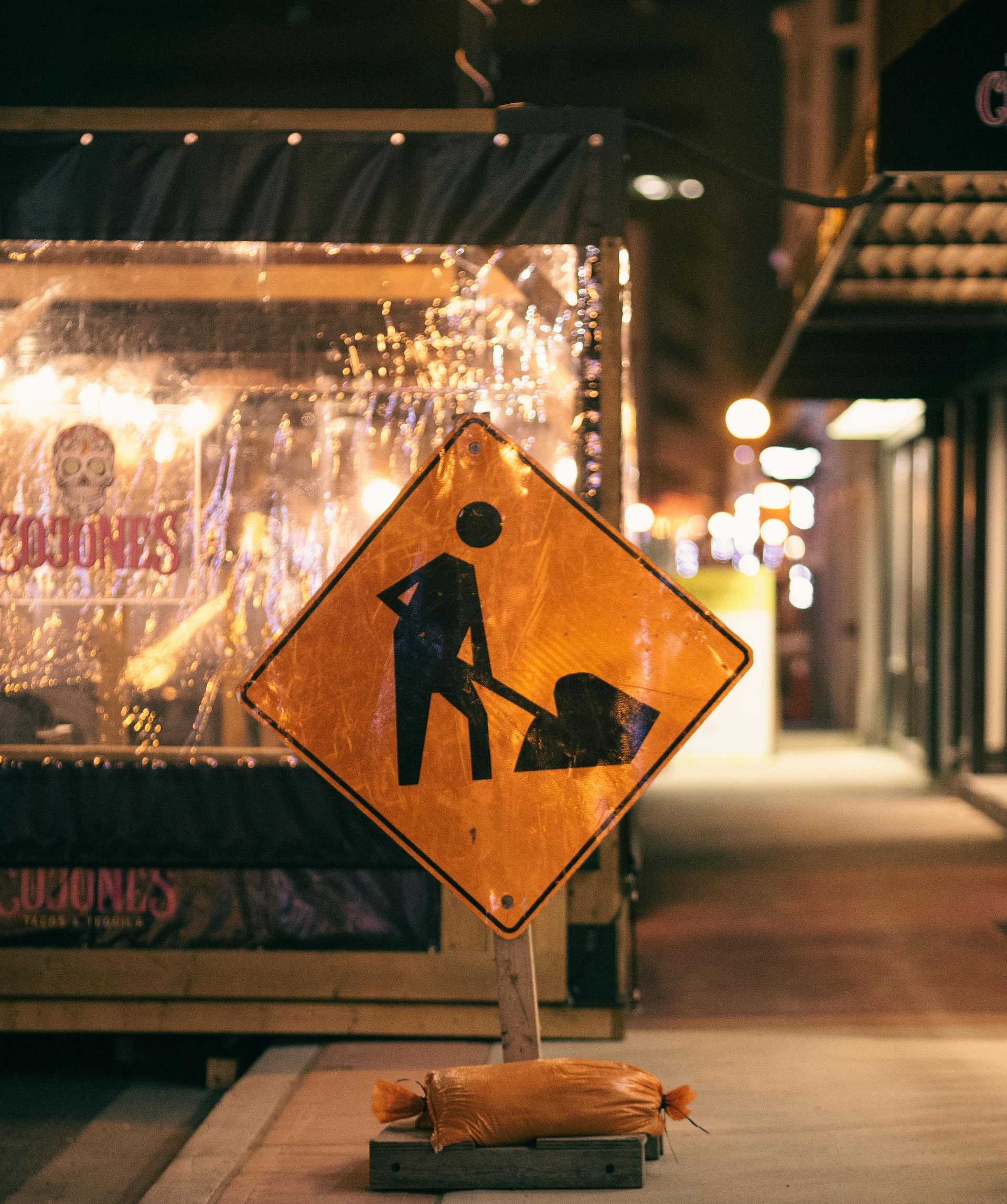 Orange construction sign on a city sidewalk at night, with storefront lights in the background