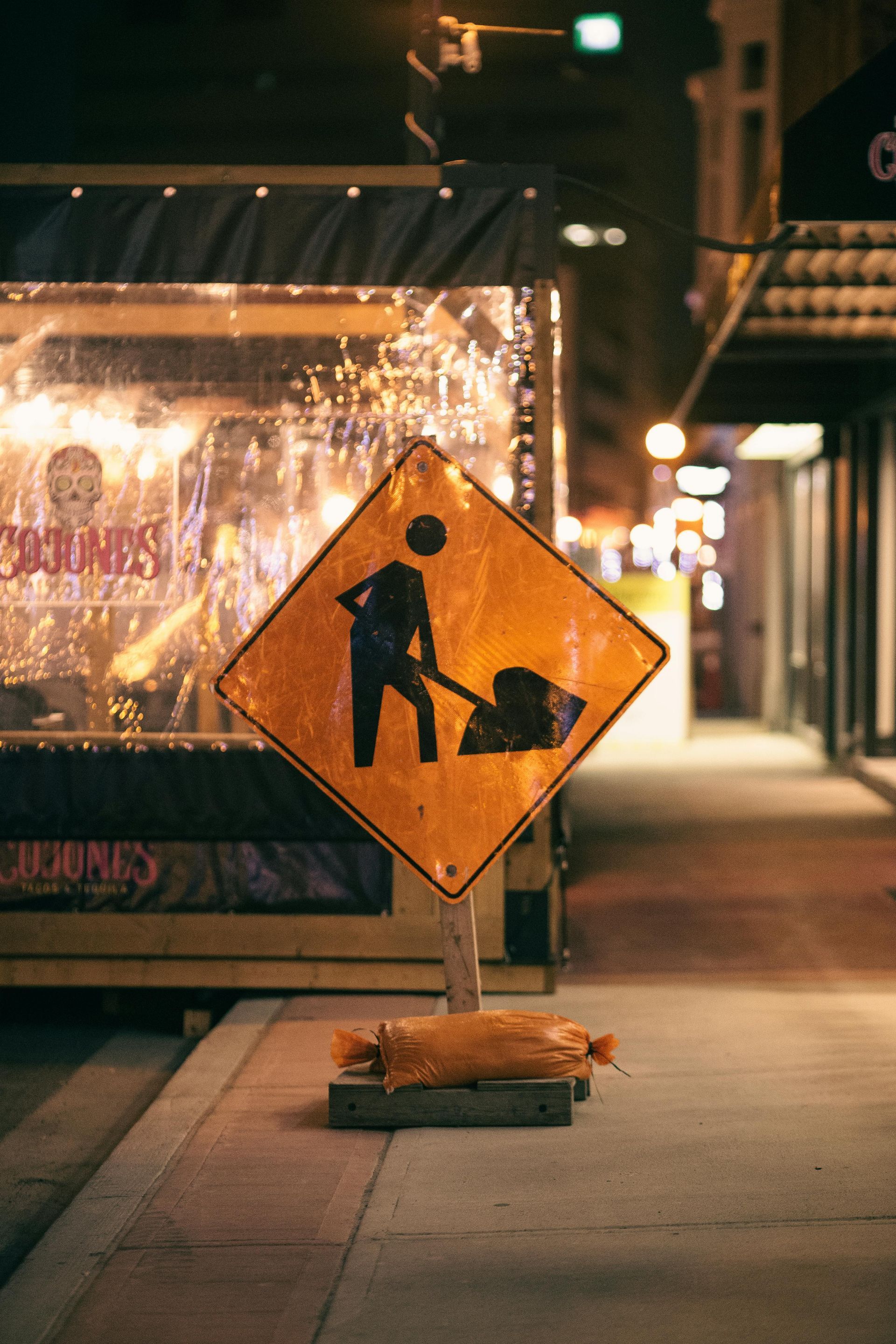 Orange construction sign on a city sidewalk at night, with storefront lights in the background