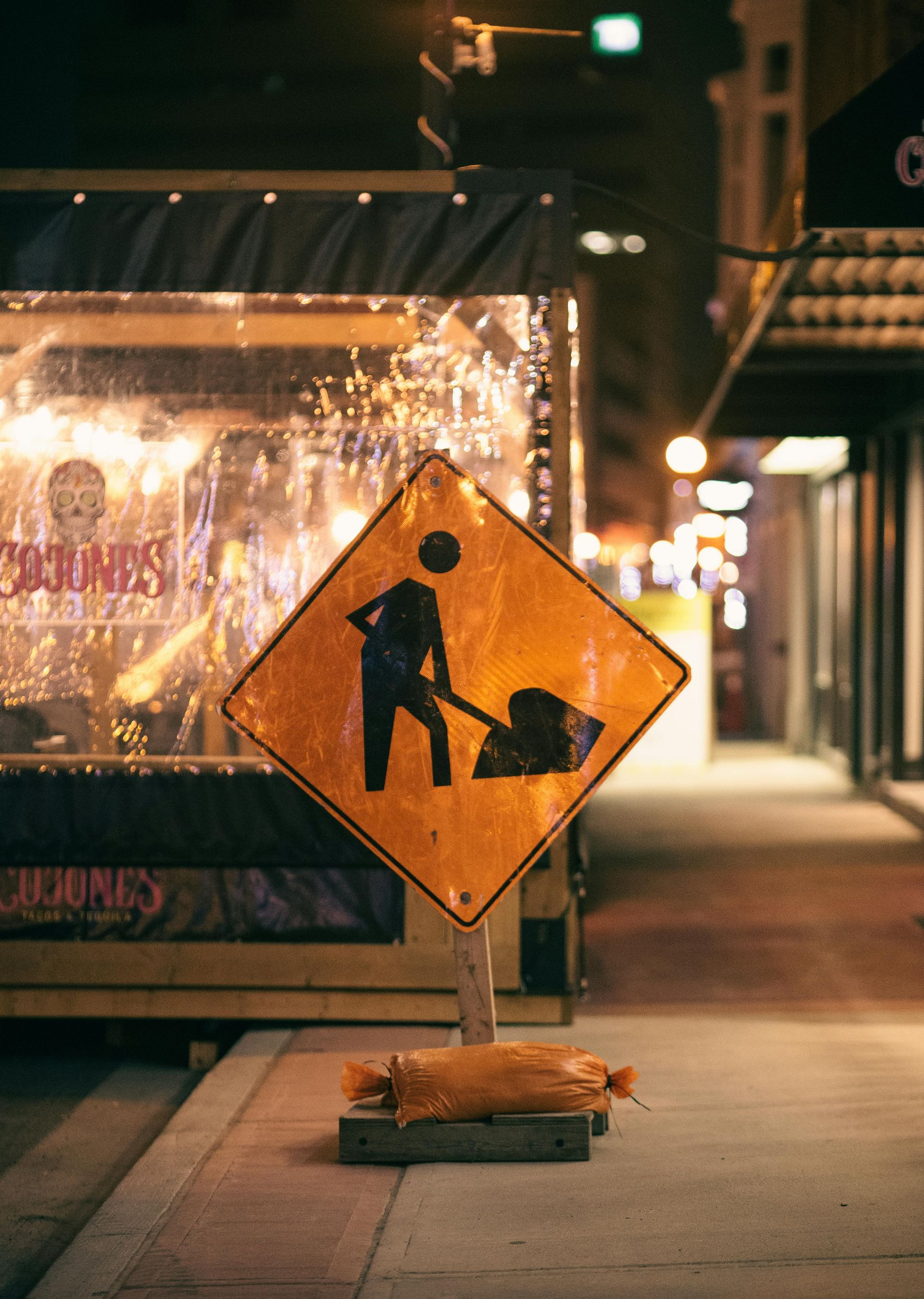 Orange construction sign on a city sidewalk at night, with storefront lights in the background