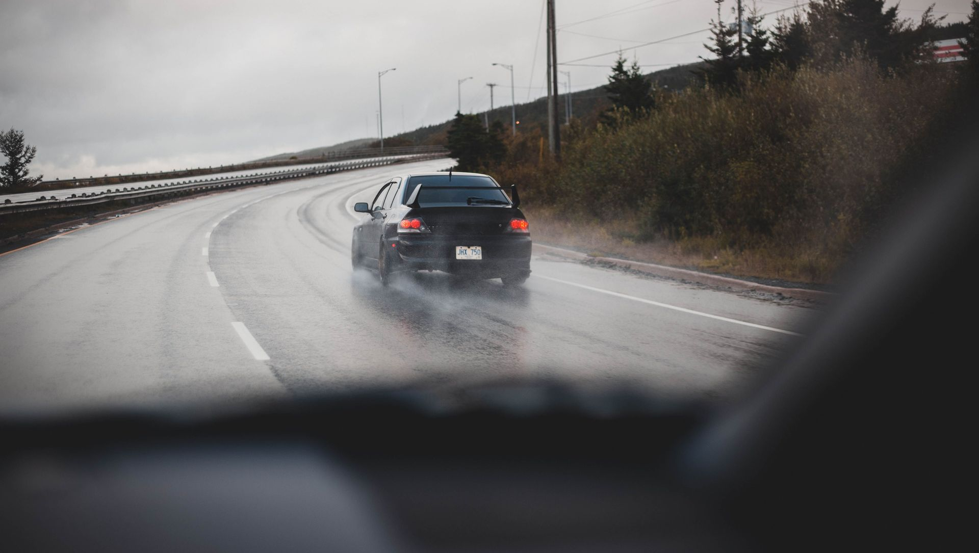 Car ahead driving on a wet, curving highway on a rainy day, seen through a car windshield.