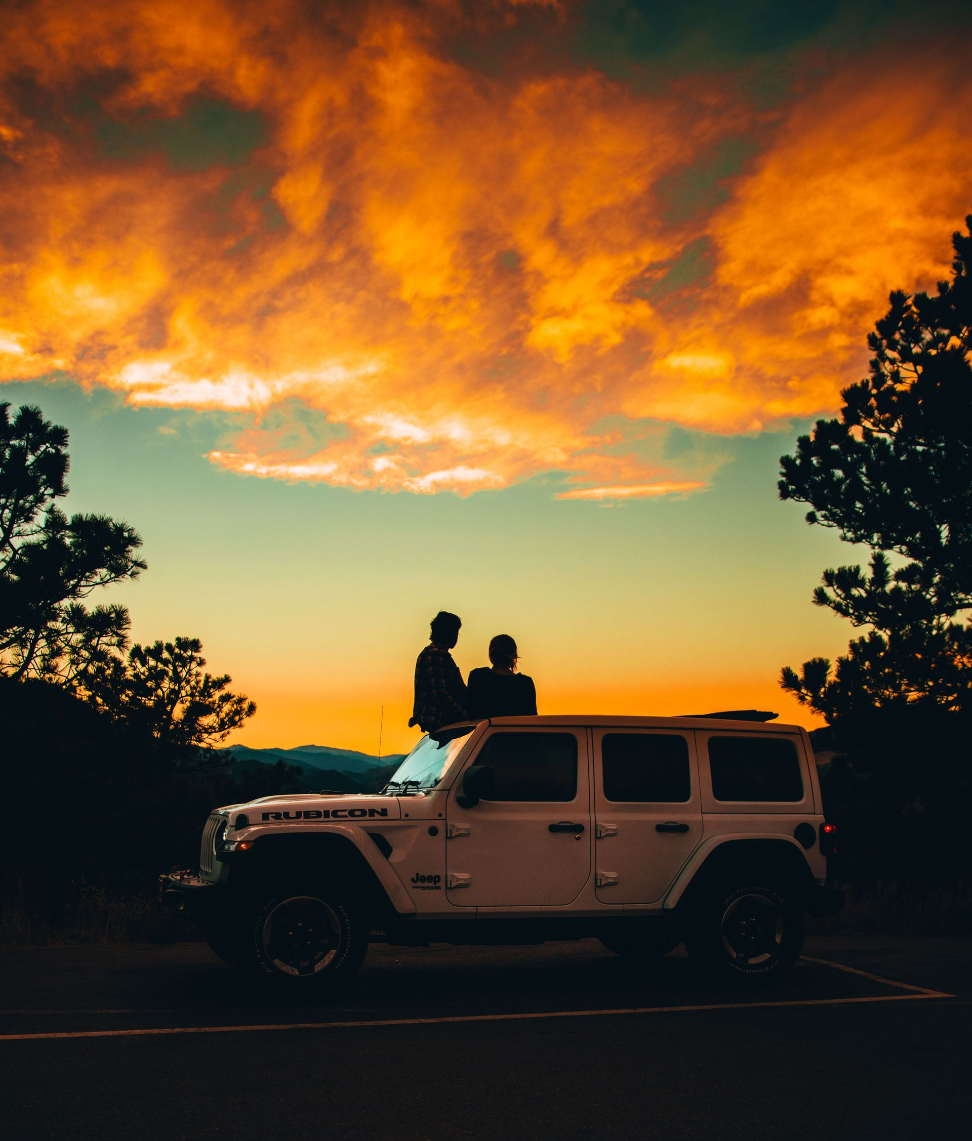 Silhouetted couple on a Jeep at sunset, under glowing orange clouds and framed by trees