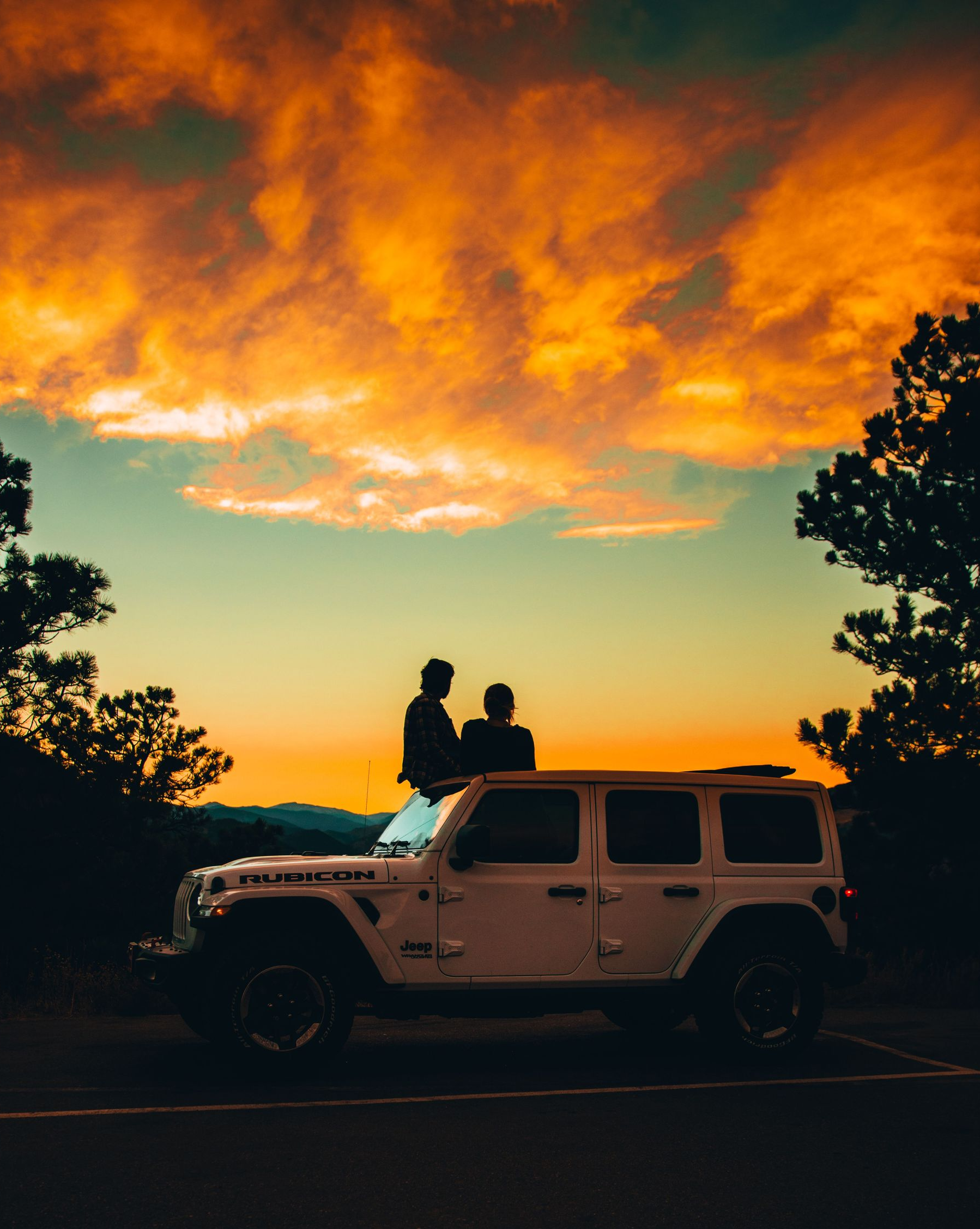Silhouetted couple on a Jeep at sunset, under glowing orange clouds and framed by trees