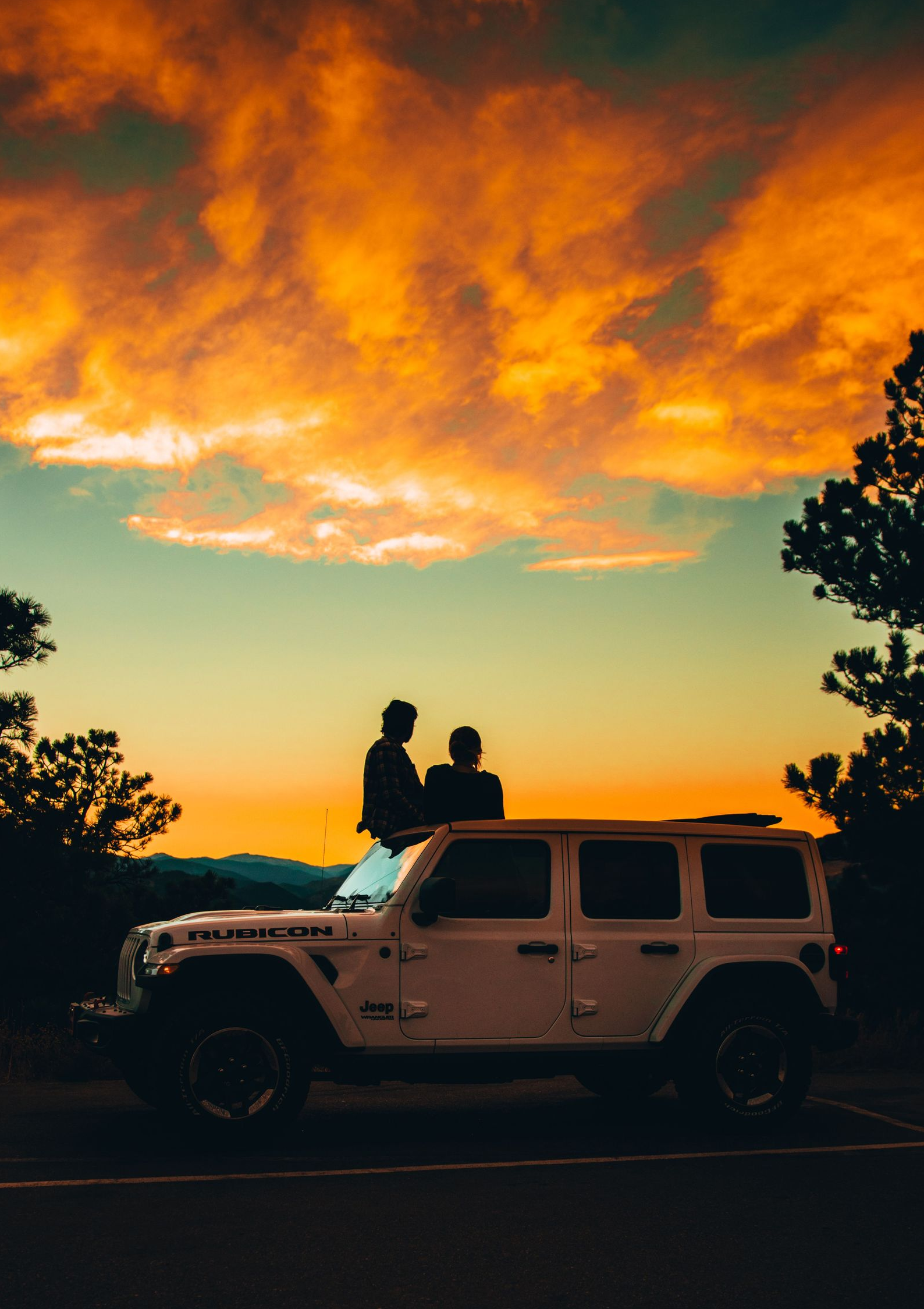Silhouetted couple on a Jeep at sunset, under glowing orange clouds and framed by trees