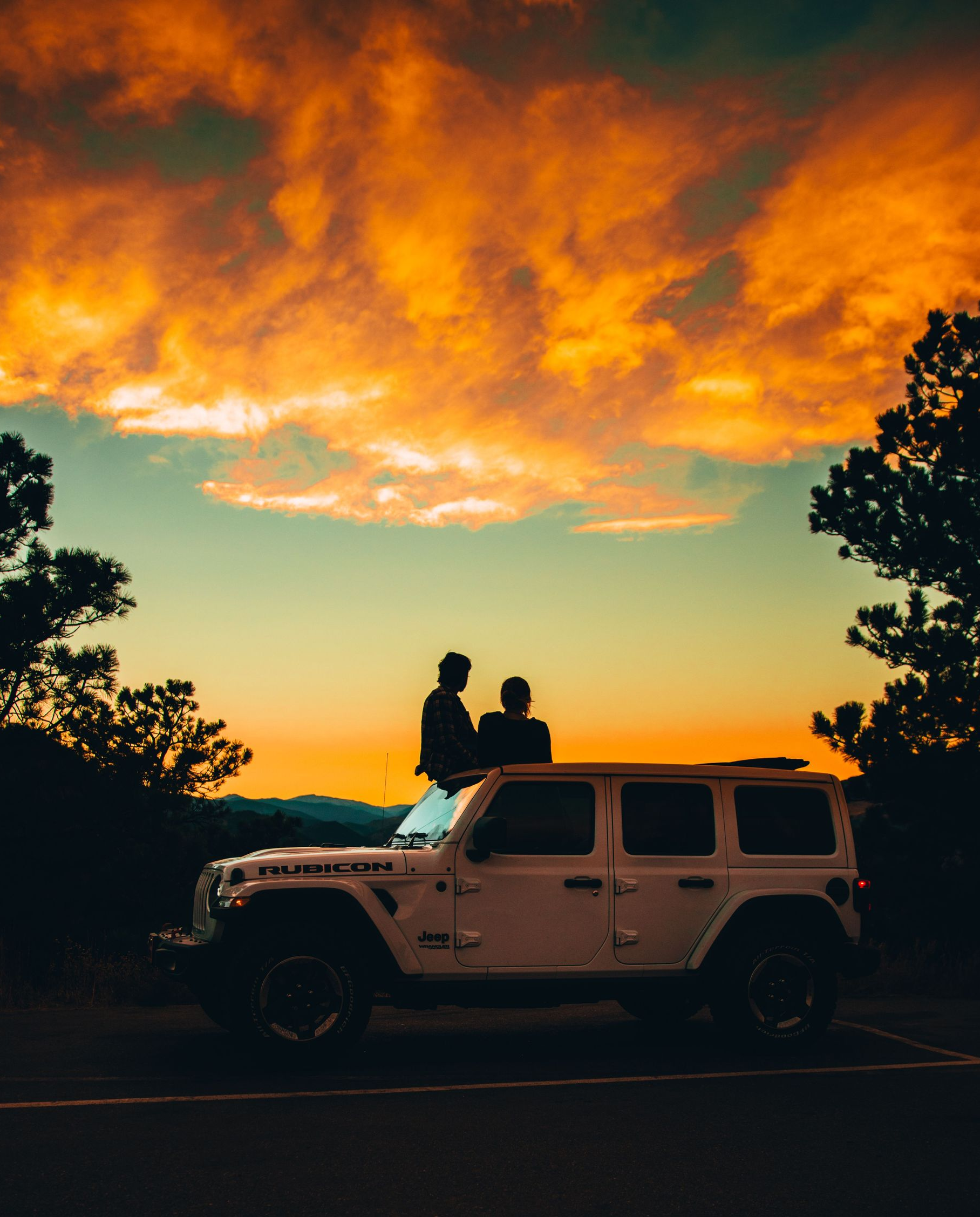Silhouetted couple on a Jeep at sunset, under glowing orange clouds and framed by trees