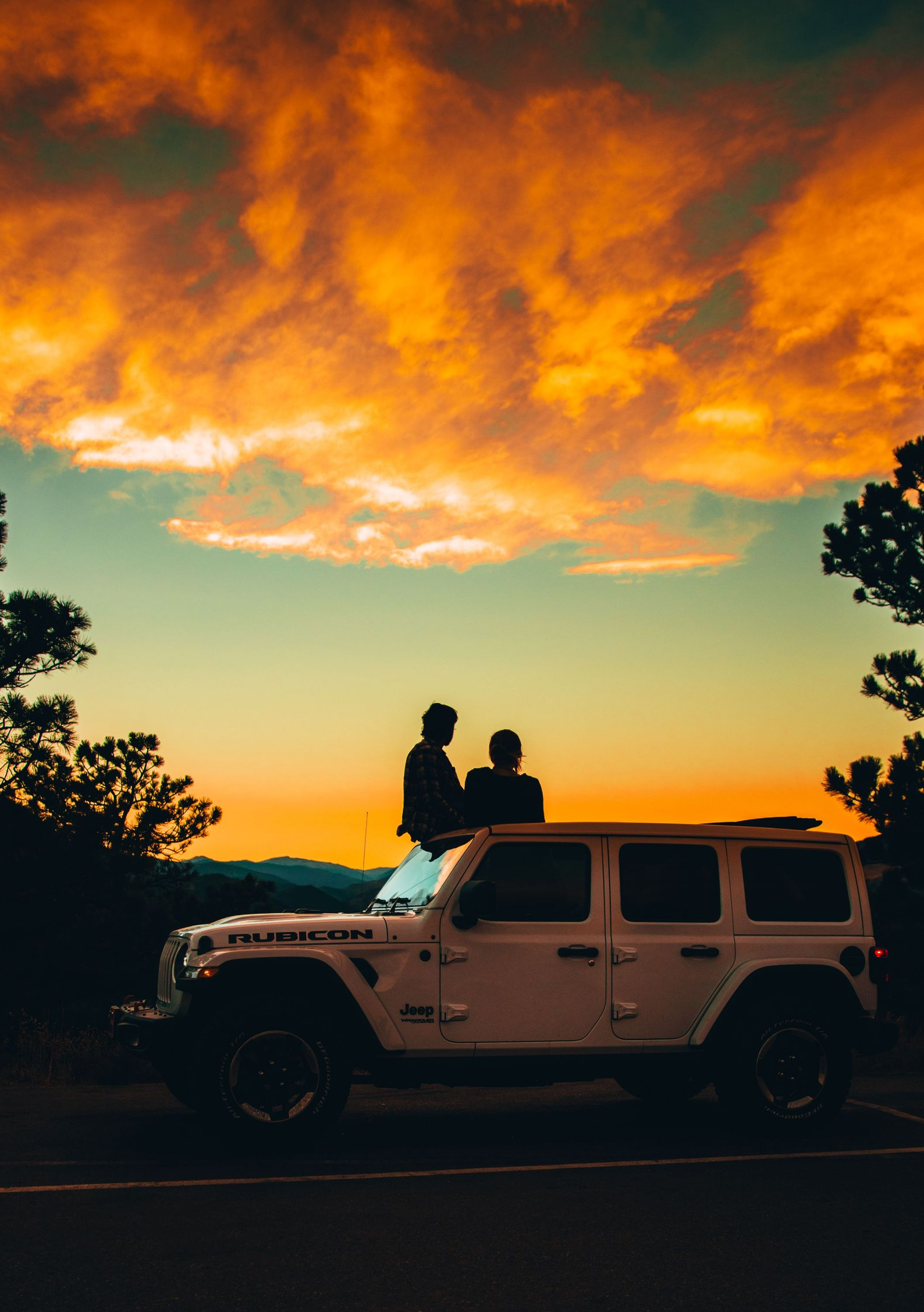 Silhouetted couple on a Jeep at sunset, under glowing orange clouds and framed by trees