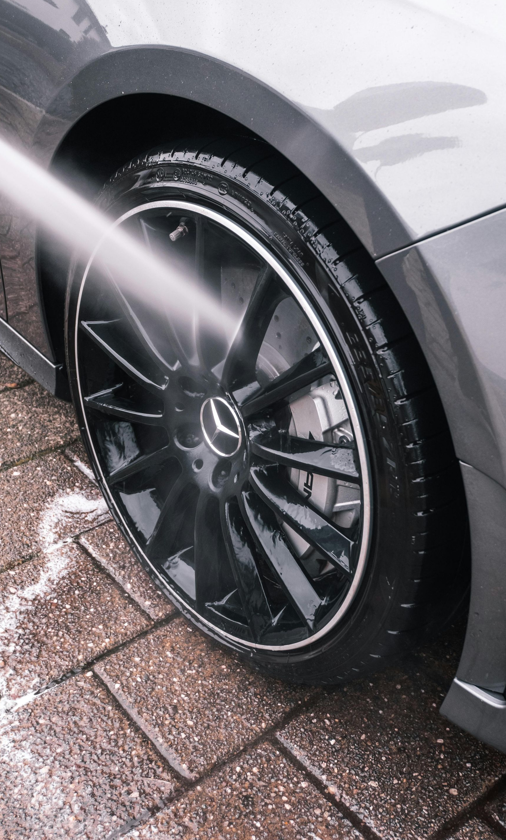 A pressure washer sprays water onto the shiny black wheel and tire of a gray Mercedes-Benz on a paved driveway.