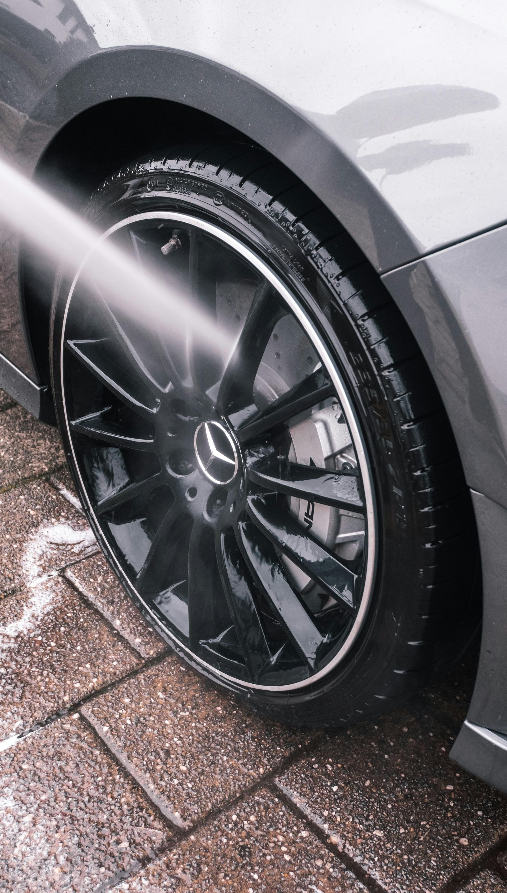 A pressure washer sprays water onto the shiny black wheel and tire of a gray Mercedes-Benz on a paved driveway.