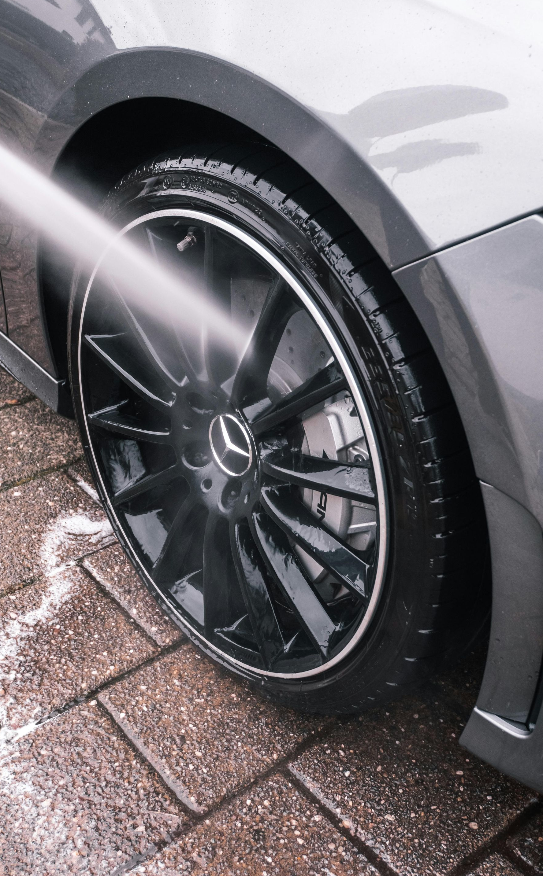 A pressure washer sprays water onto the shiny black wheel and tire of a gray Mercedes-Benz on a paved driveway.