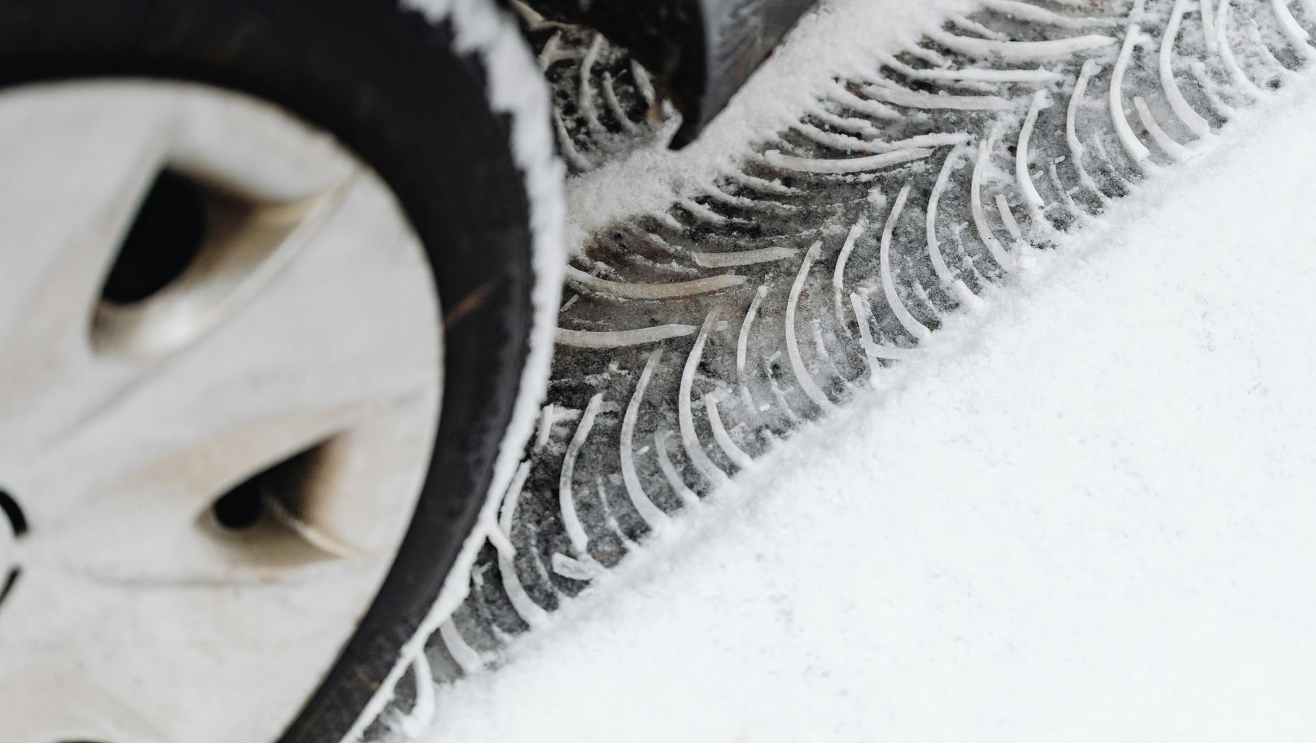 A car tire with a deep, directional tread pattern driving through fresh, white snow, leaving a clear track behind it.