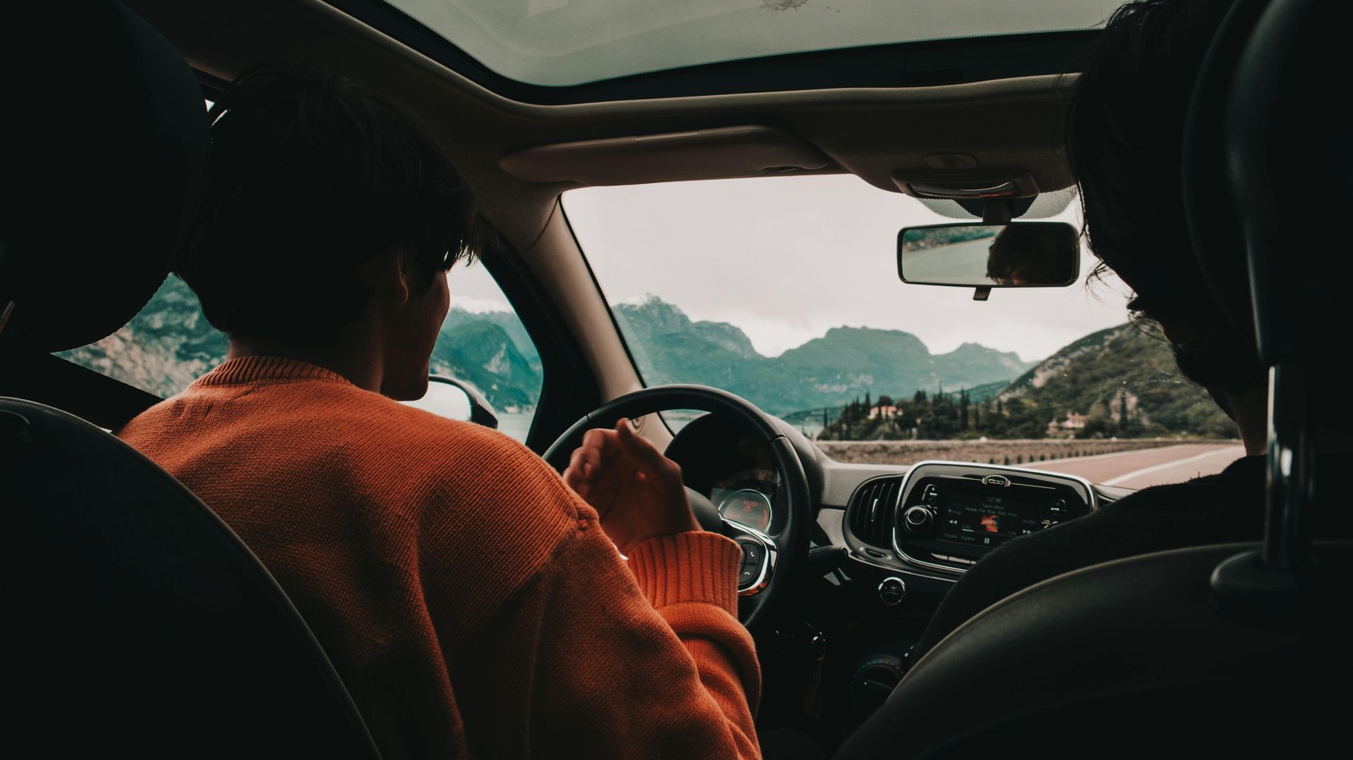 Person in orange sweater drives a car with a passenger, mountains visible through the windshield.