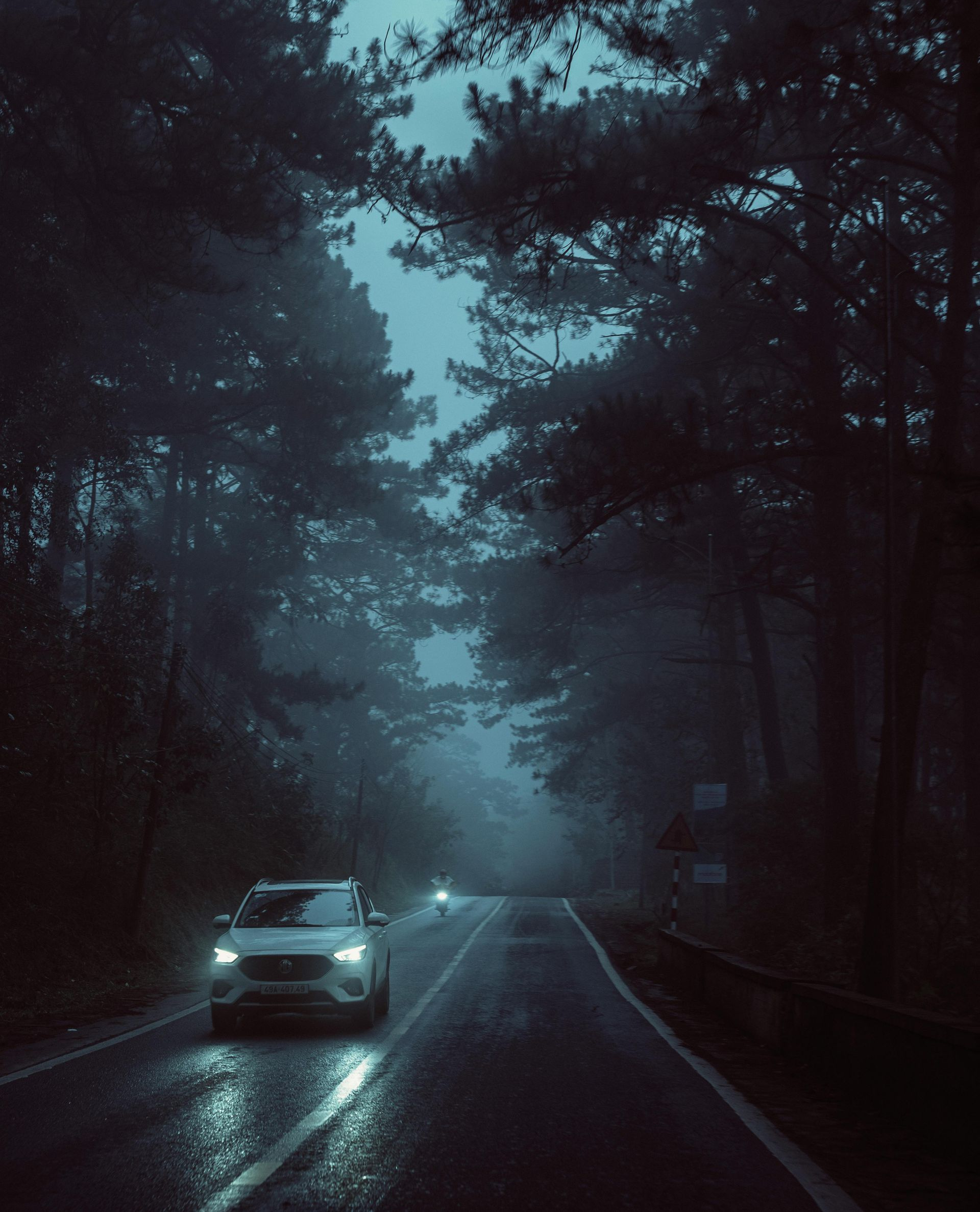 Car driving on a foggy, dark road through a forest at night. Headlights illuminate the wet asphalt.