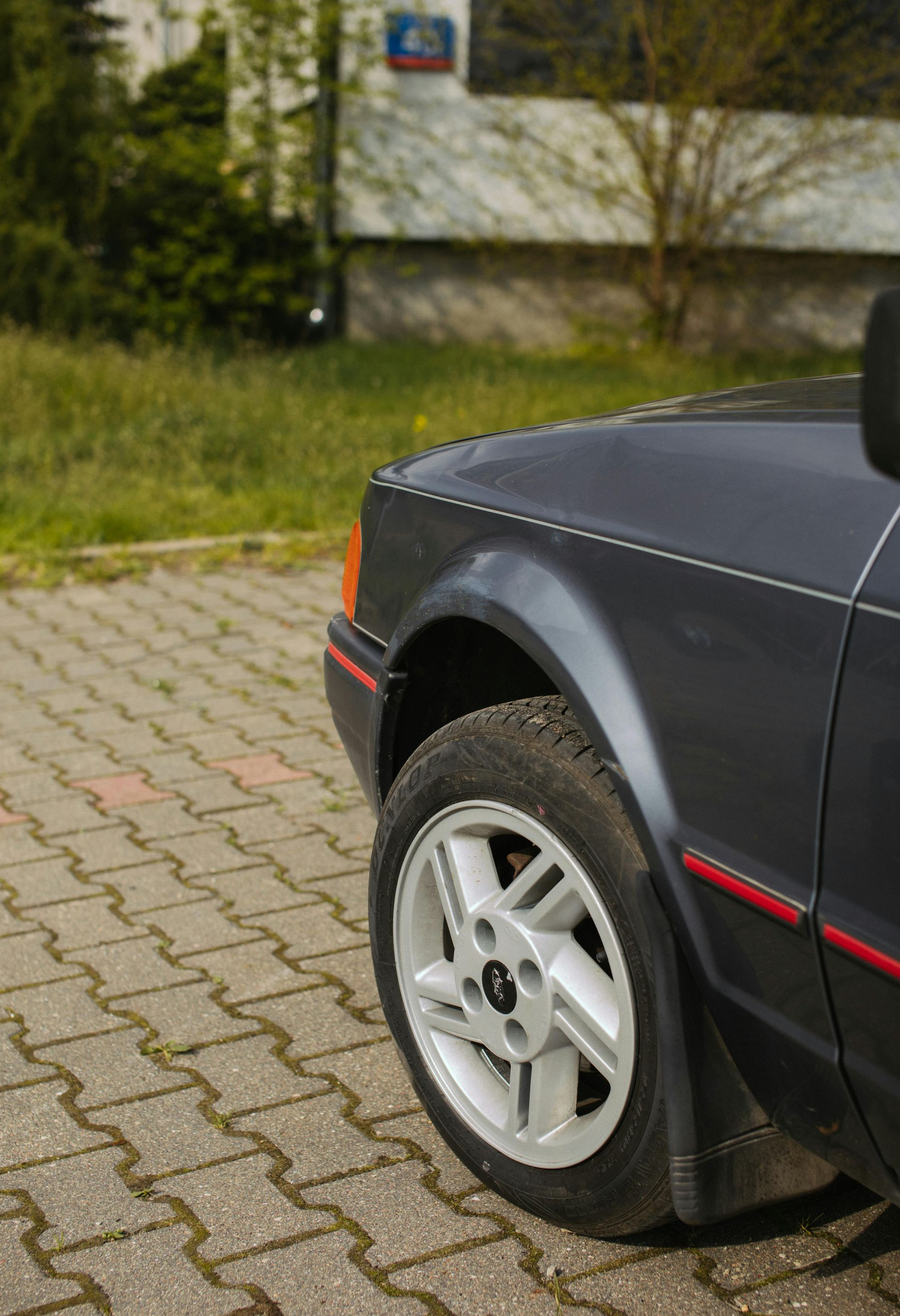 Dark gray Ford car's front quarter panel and wheel. Gray wheels, red trim, on a paved surface.