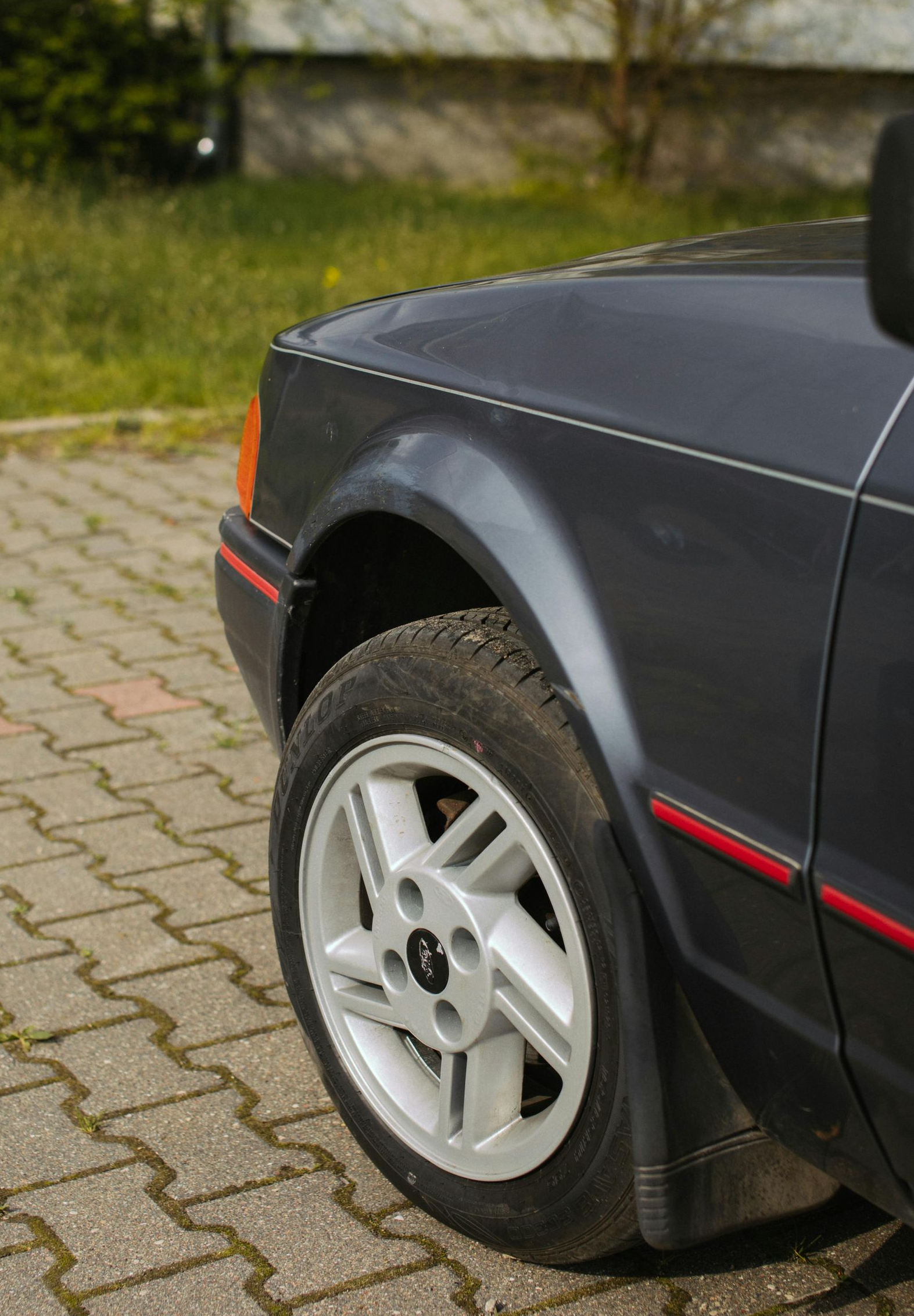 Gray car's front quarter, showing wheel, fender, and part of the hood. Red trim detail. Parked on paving stones.