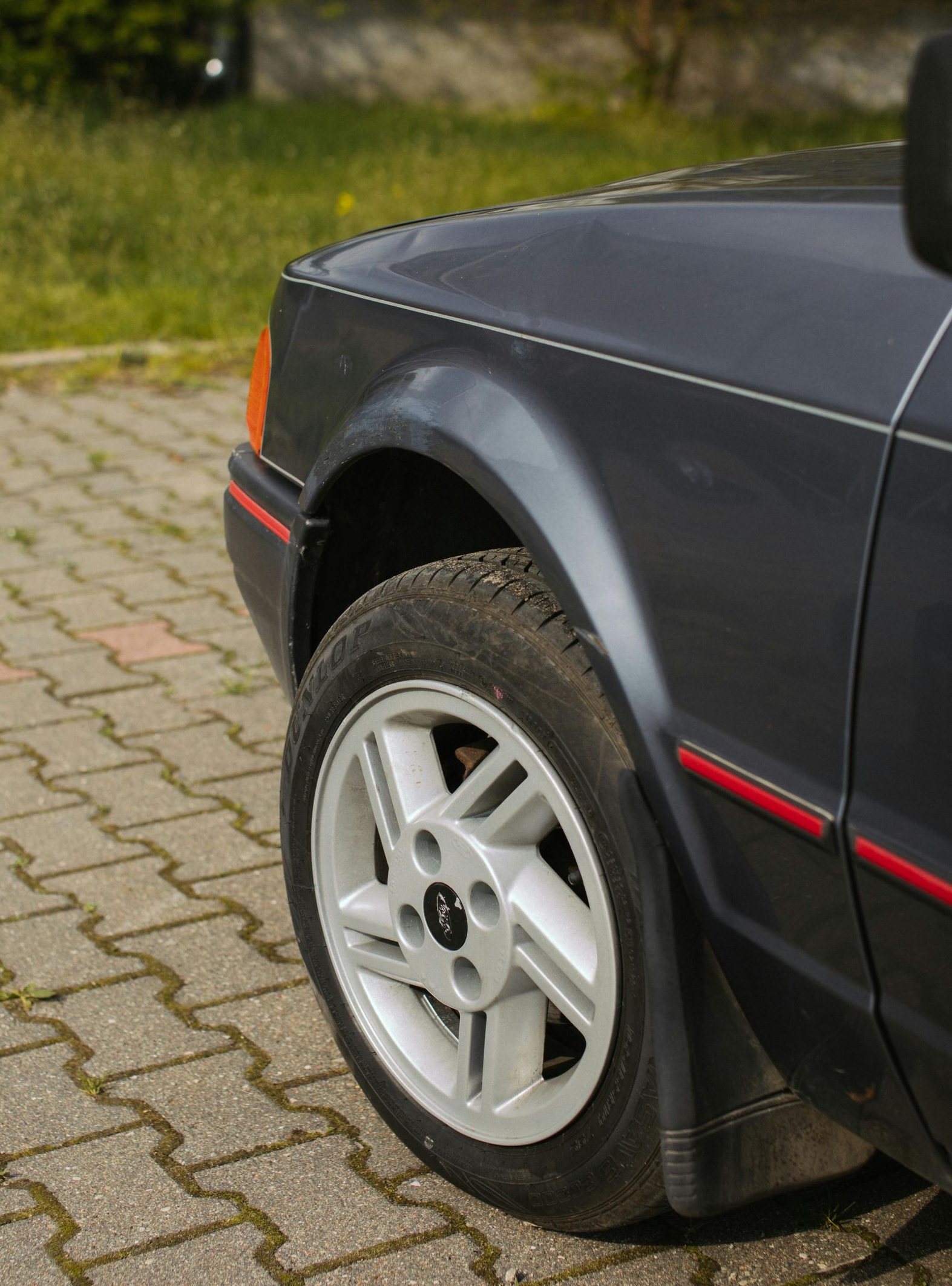 Gray car's front quarter, showing wheel, fender, and part of the hood. Red trim detail. Parked on paving stones.