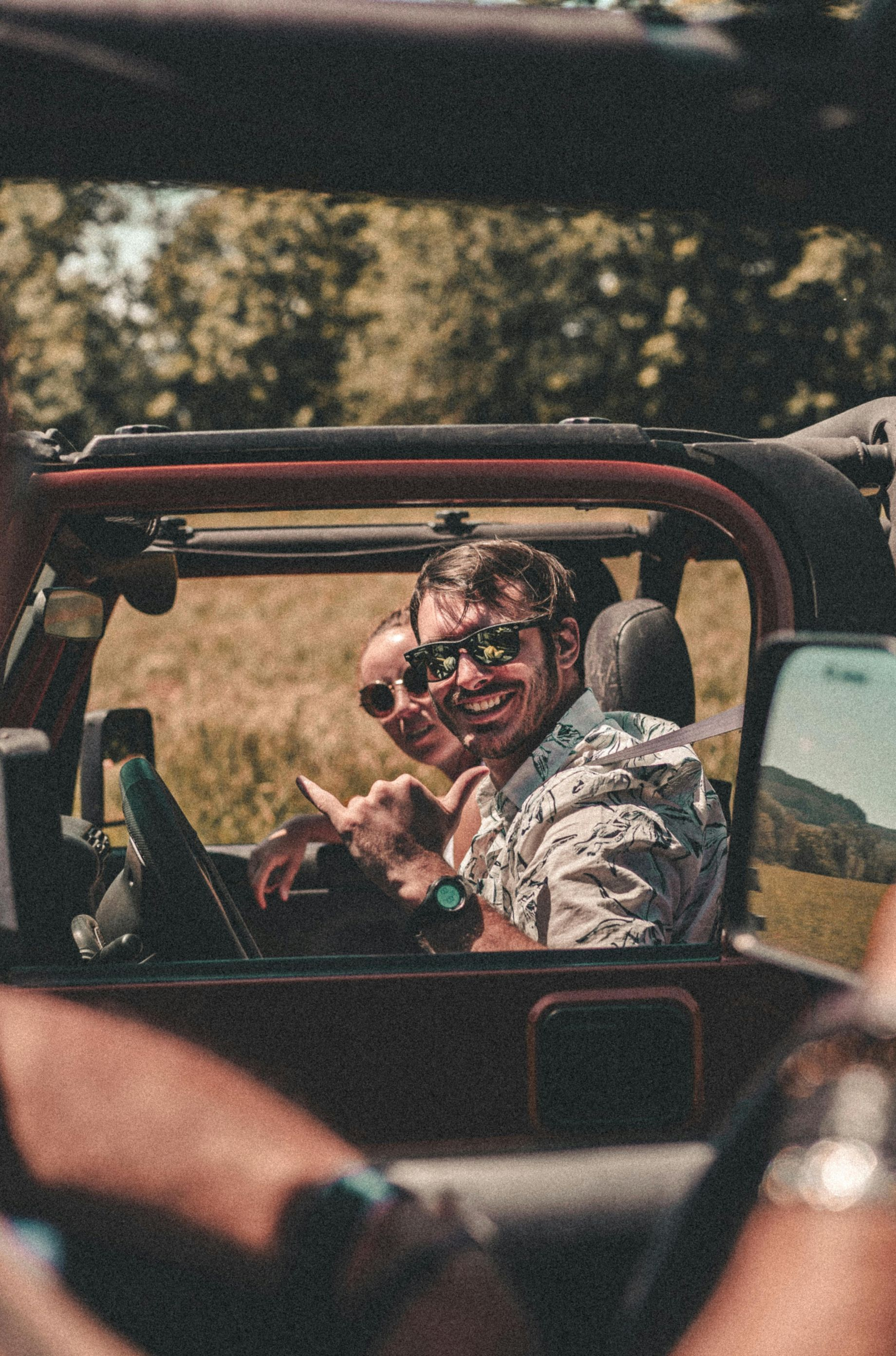 Two people smiling in a red open-top vehicle on a sunny road, with another passenger in the foreground
