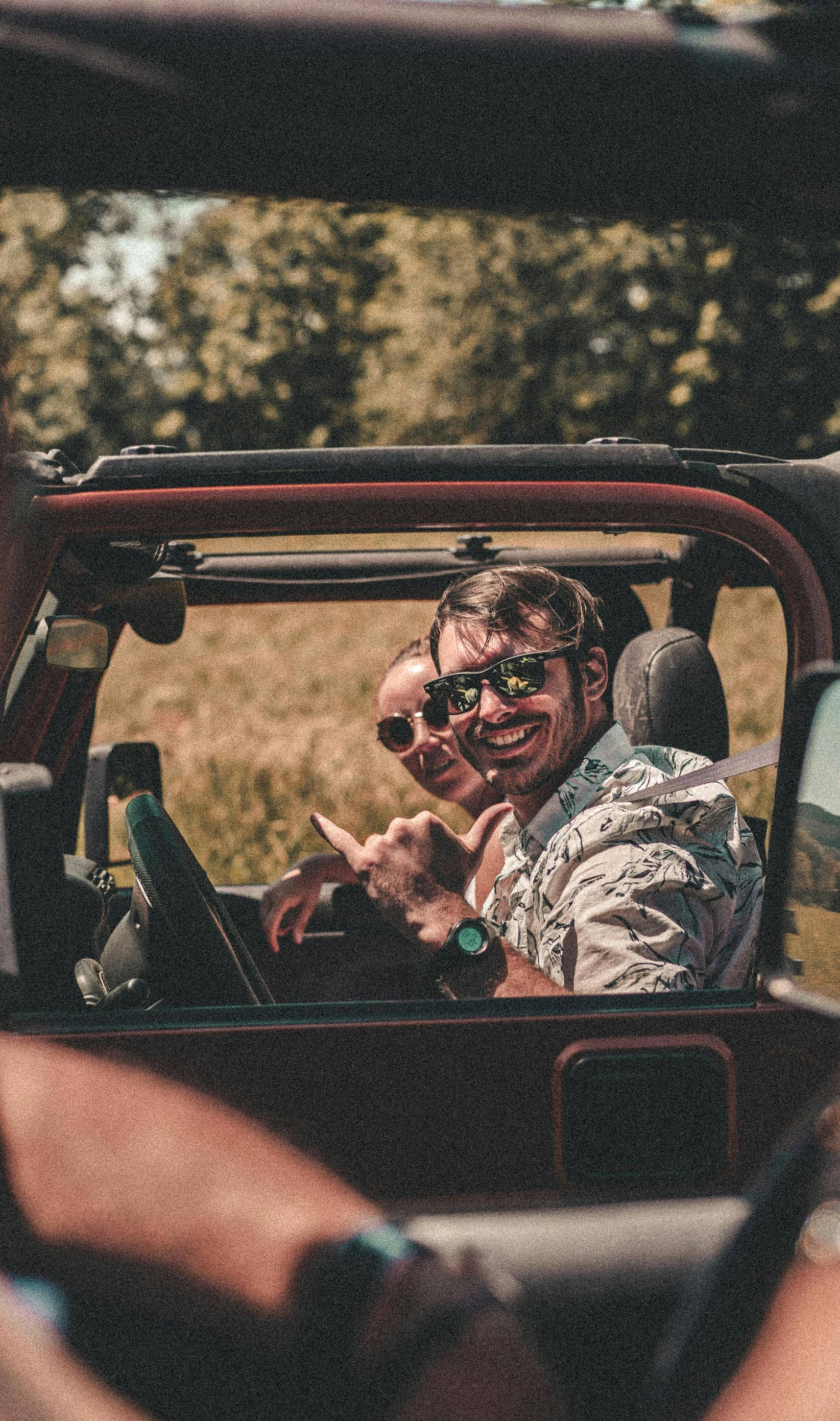 Two people smiling in a red open-top vehicle on a sunny road, with another passenger in the foreground