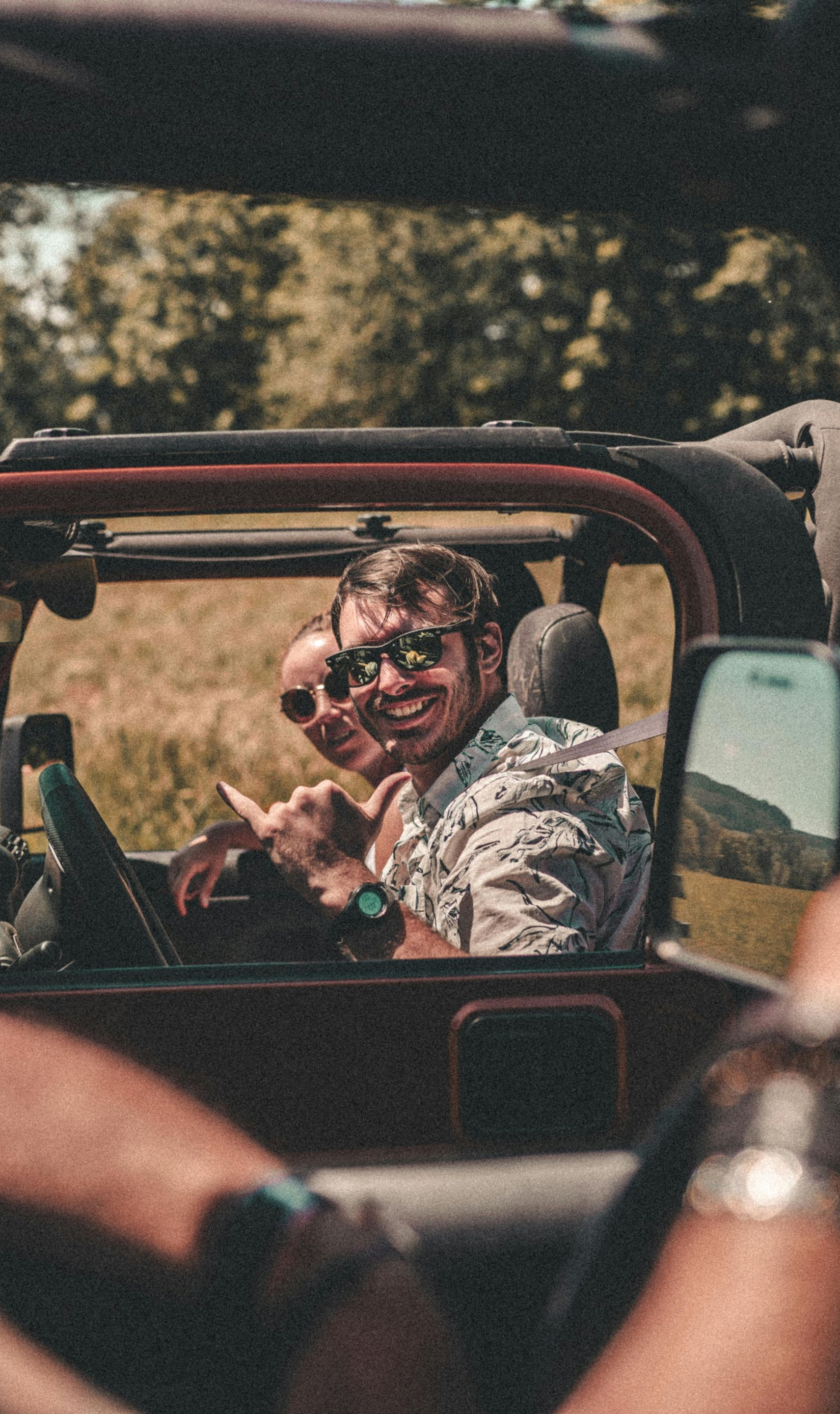 Two people smiling in a red open-top vehicle on a sunny road, with another passenger in the foreground