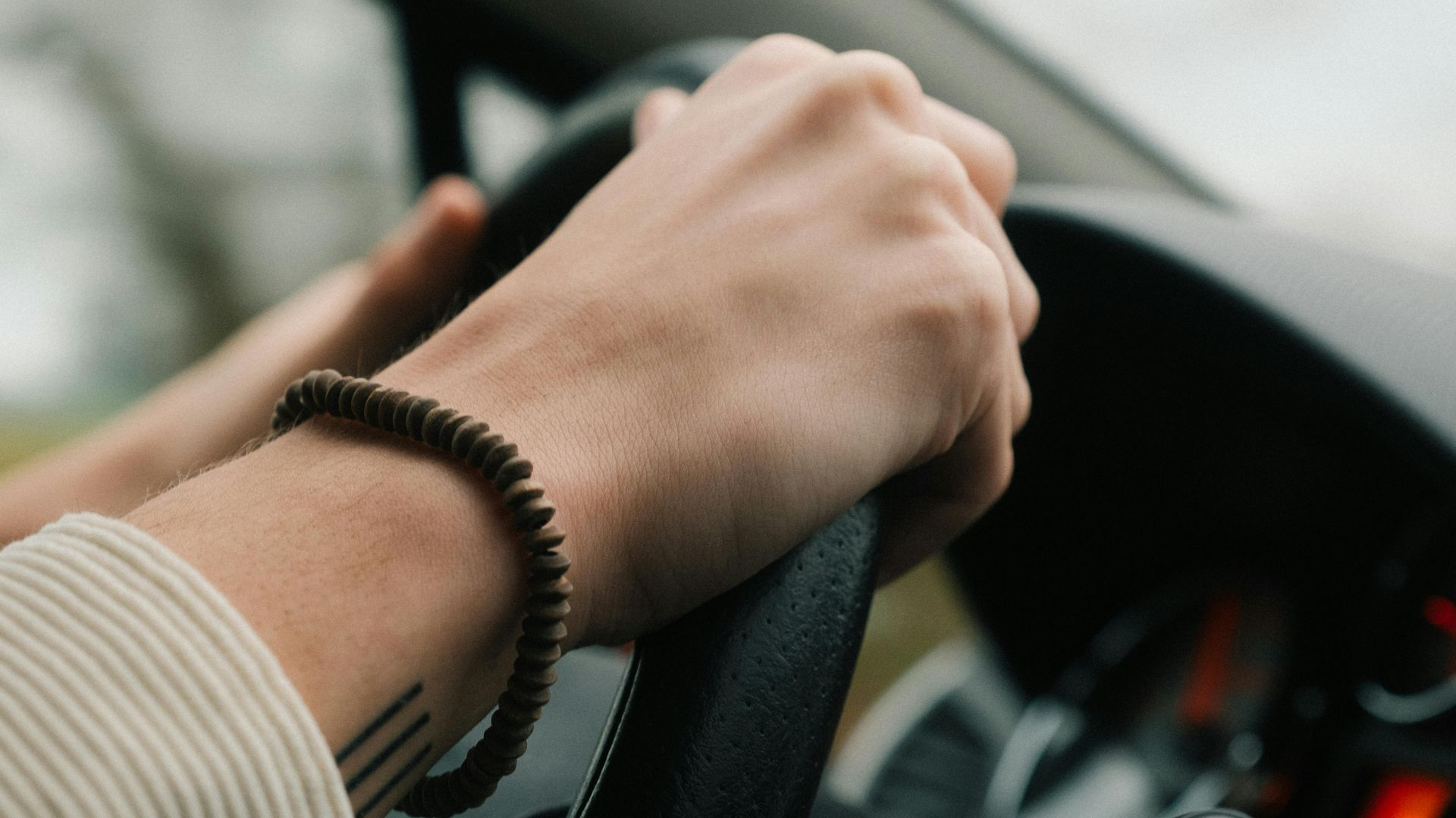 Hands gripping a steering wheel inside a car, with a beaded bracelet visible.