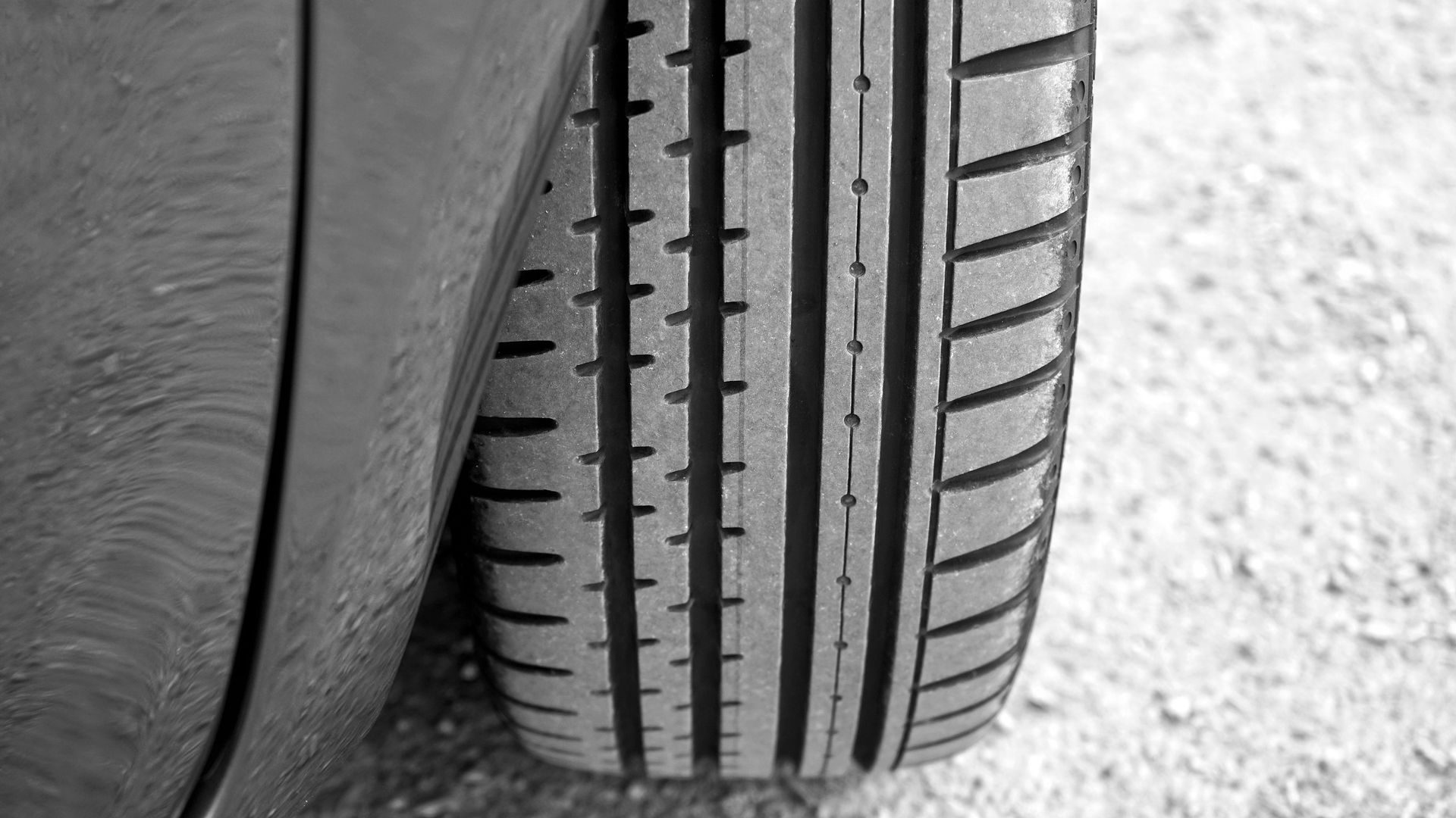 Black and white close-up of a car tire with tread lines on a gravel surface.