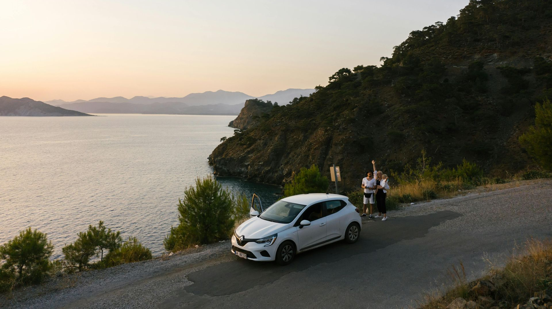 White car and cyclists on a coastal road at sunset, with calm water and hills in the background