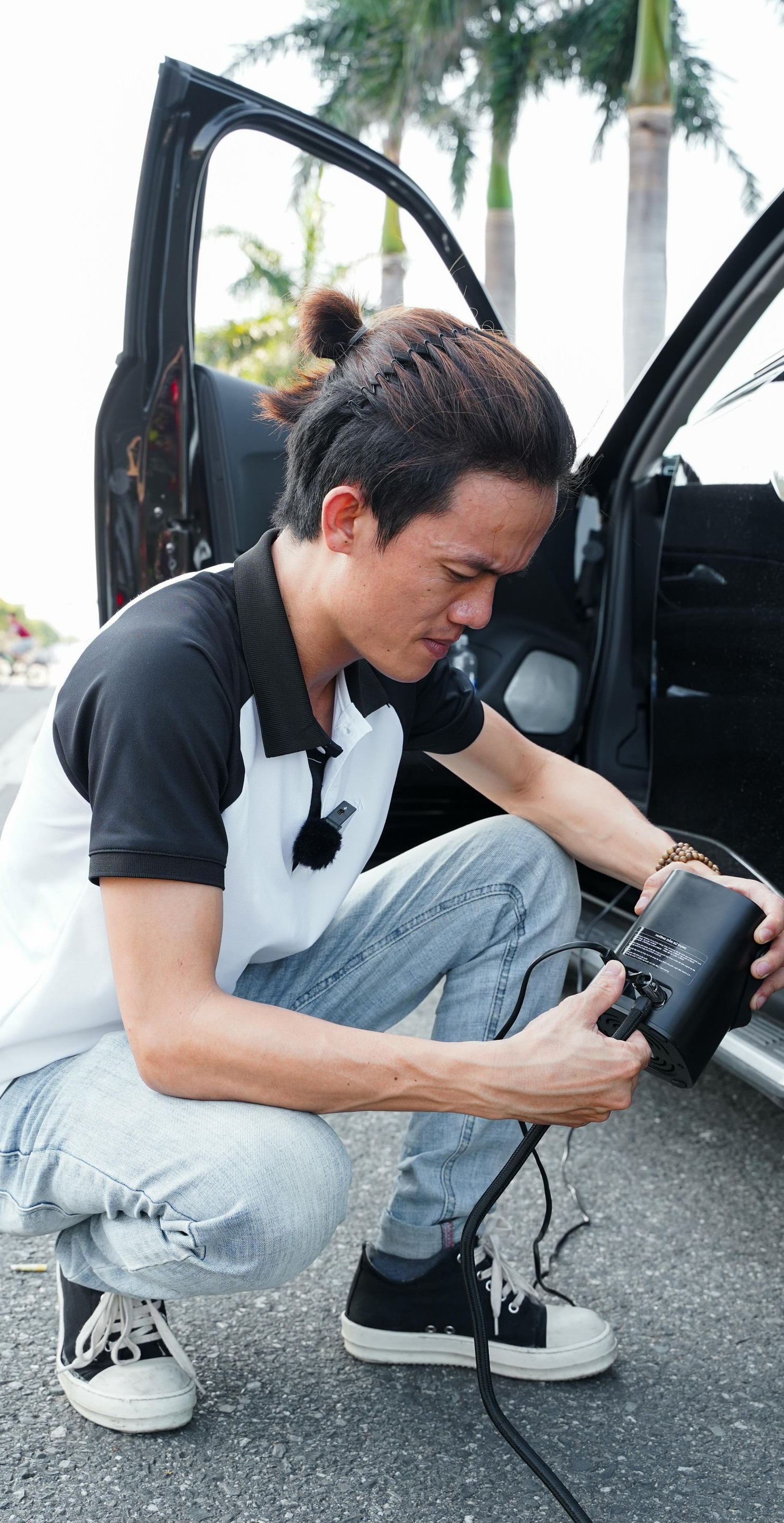 Man crouches next to a car, connecting a black cable. Open car door, outdoor setting.