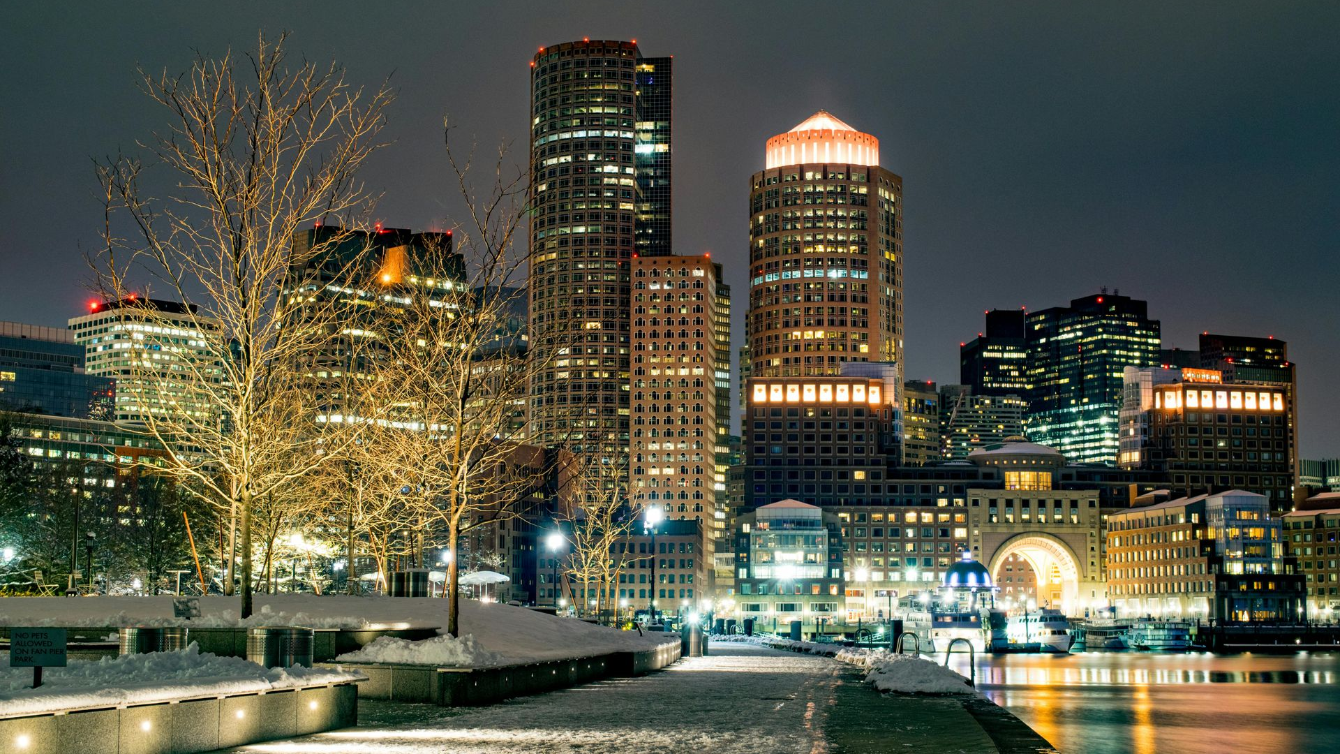 Boston skyline at night, illuminated buildings reflected in harbor; snow-covered walkway with lit trees.